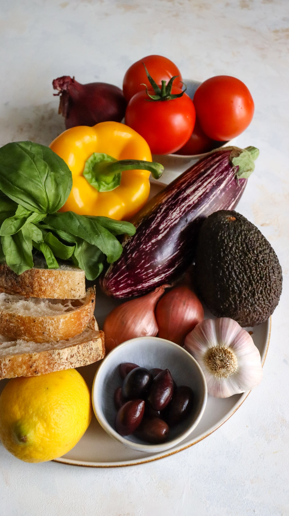 Teller mit Tomaten, Paprika, Aubergine, Avocado, Brot, Zitronen, Basilikum, Oliven, Zwiebeln und Knoblauch.