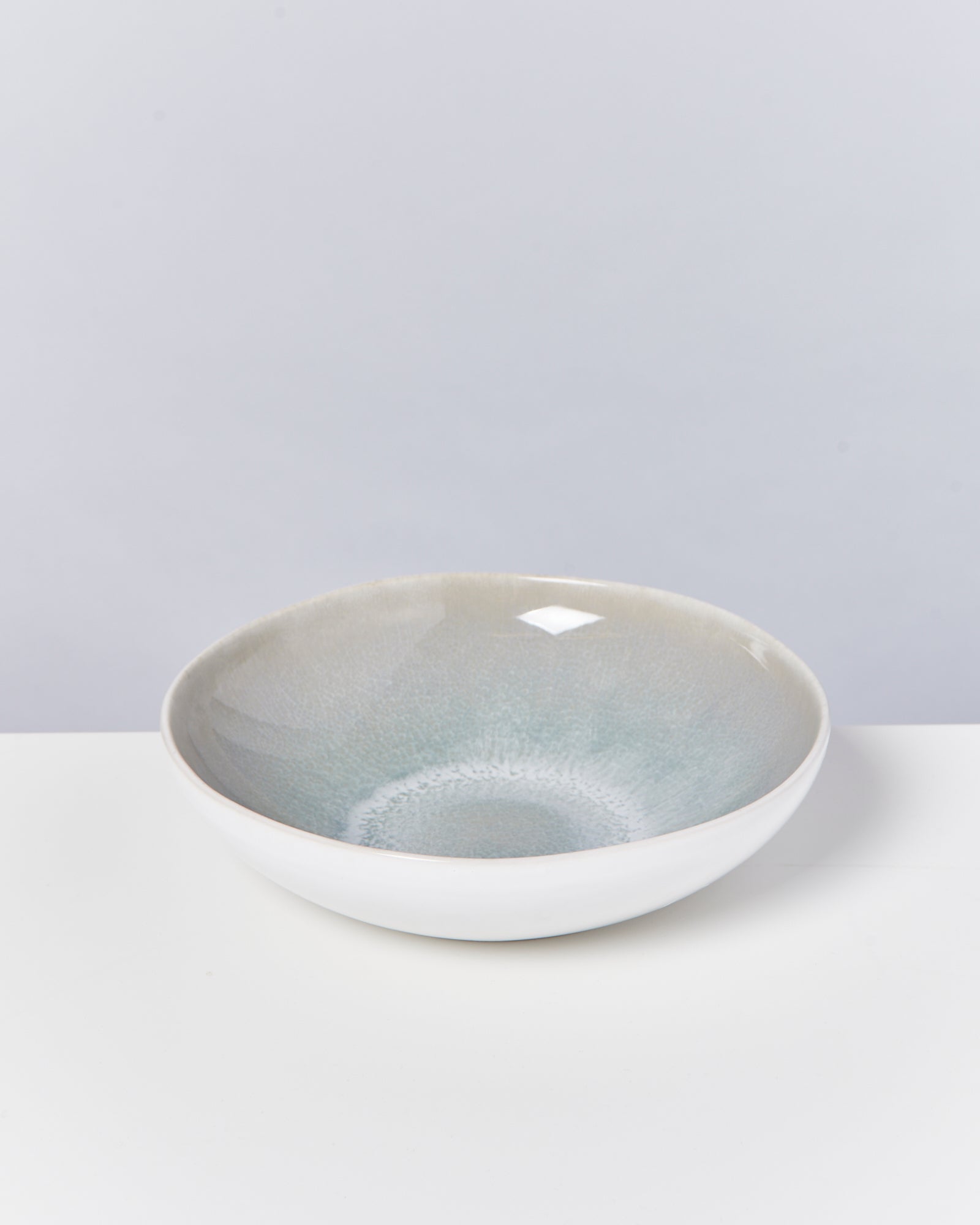 Eine graue, flache Schüssel mit sanftem blauem Schimmer steht auf einem weißen Tisch vor grauem Hintergrund.