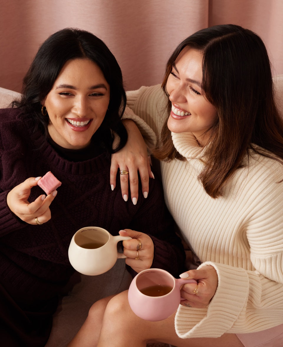 Zwei lachende Frauen sitzen mit Tassen und Gebäck in der Hand auf einem Sofa vor einem rosa Hintergrund.