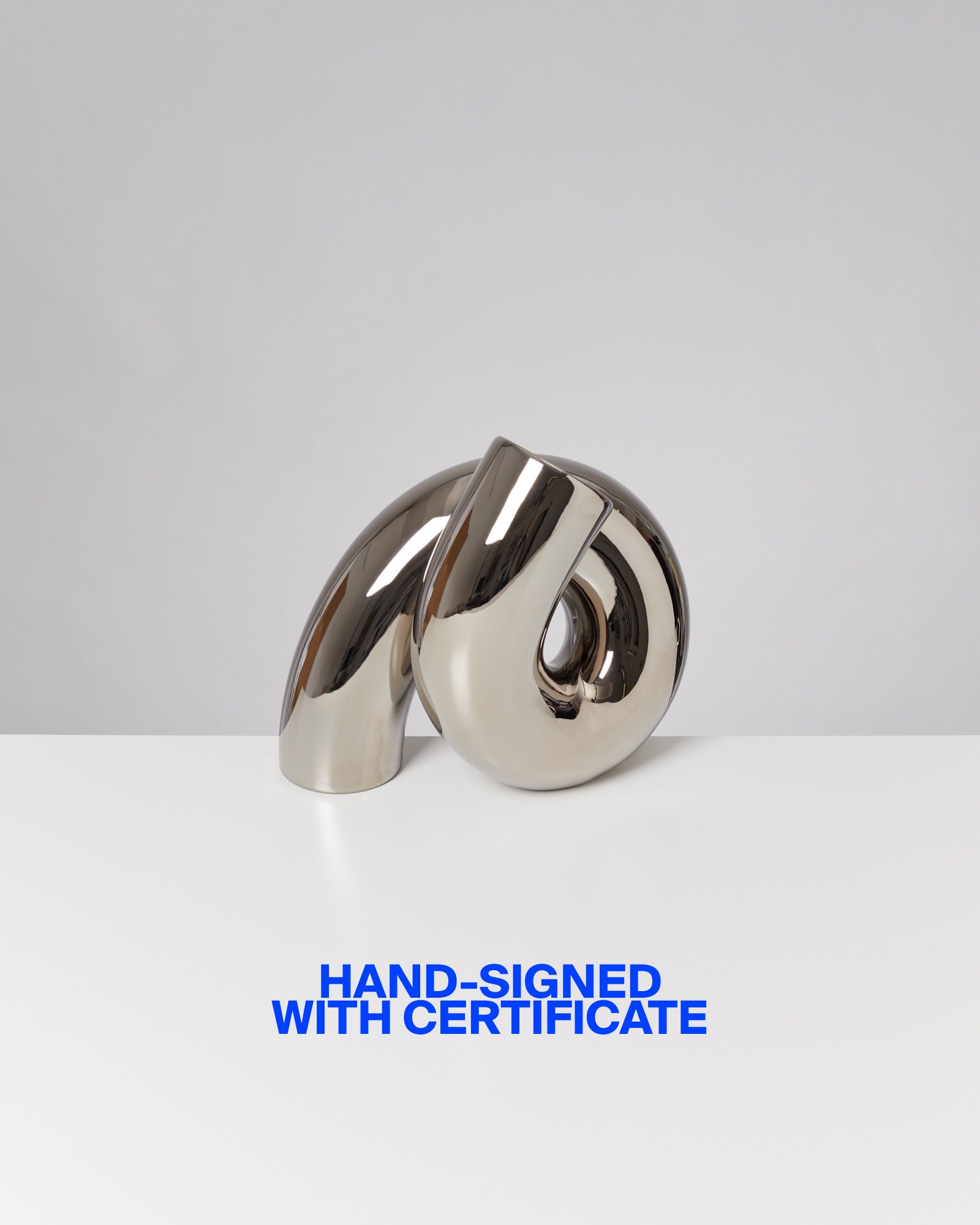 Moderne, reflektierende Skulptur auf weißem Tisch mit blauem Text: "HAND-SIGNED WITH CERTIFICATE" im Vordergrund.