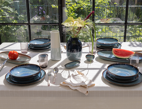 Ein gedeckter Tisch in der Sonne mit blauem Geschirr, Gläsern, einer Vase mit Blumen und Obststücken.