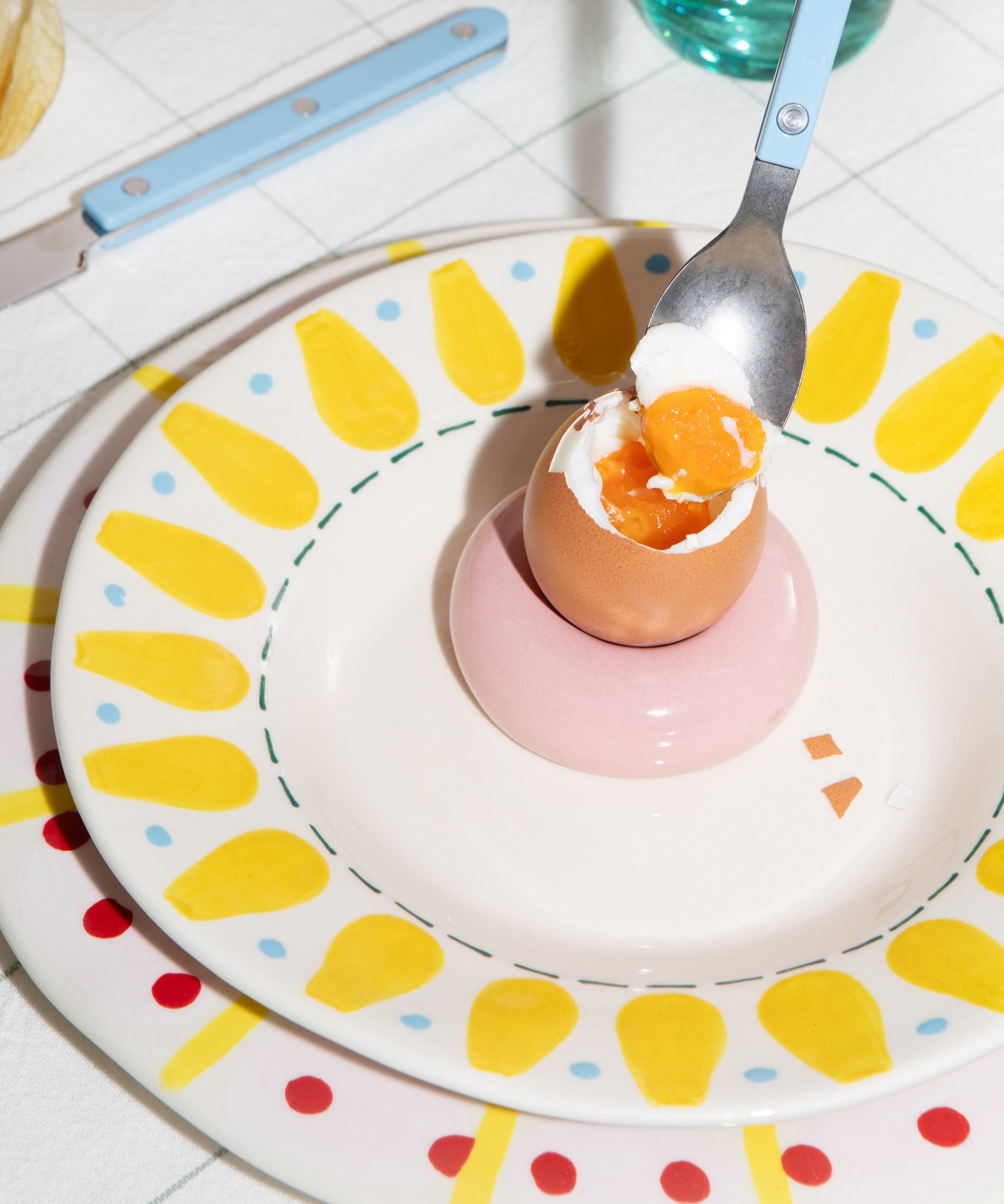 Ein gekochtes Ei in einem Eierbecher auf einem Teller mit gelbem Muster, daneben ein Löffel mit blauem Griff.