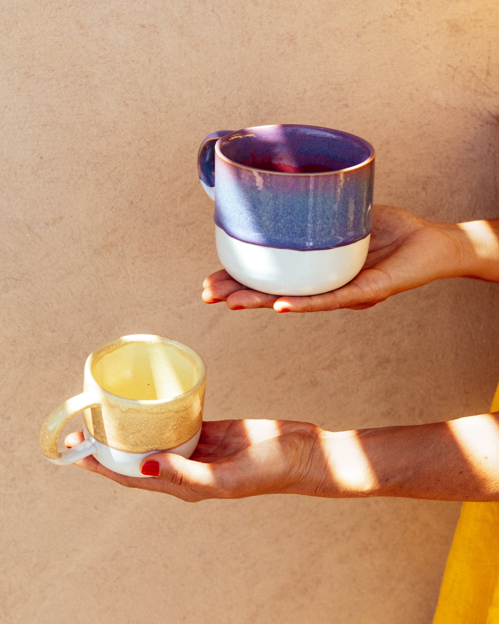 Zwei Hände halten bunte Keramiktassen, eine blau-violette und eine gelb-weiße, vor einer beigefarbenen Wand.