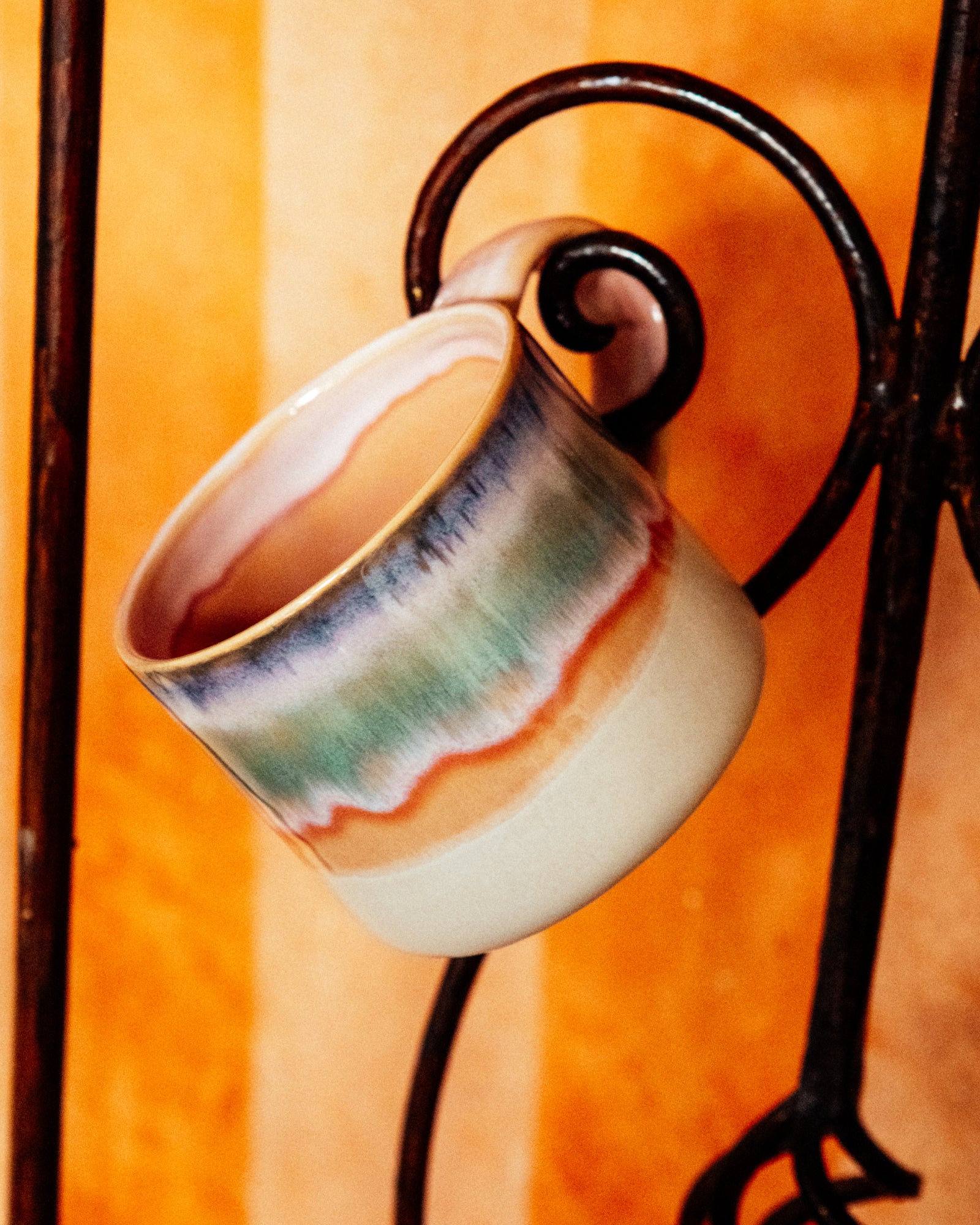 Keramiktasse mit buntem Streifenmuster hängt an schwarzem Metallhaken vor orangefarbenem Hintergrund.