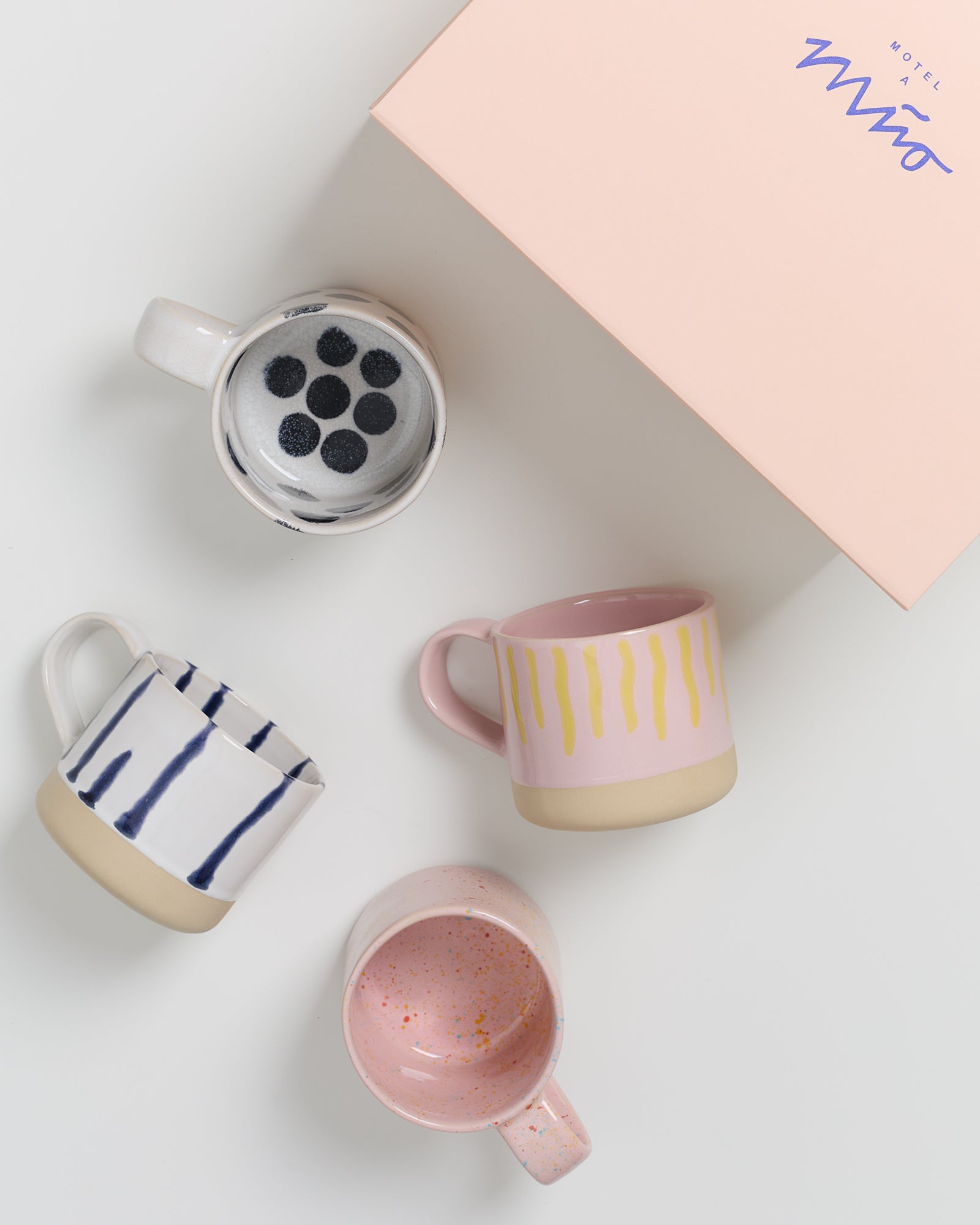 Vier bunte Keramiktassen neben einem rosa Karton auf einem weißen Hintergrund.