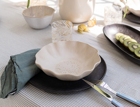 Ein gedeckter Tisch mit einer beigefarbenen Schale auf einem schwarzen Teller, daneben ein Messer, Löffel und grüne Serviette.