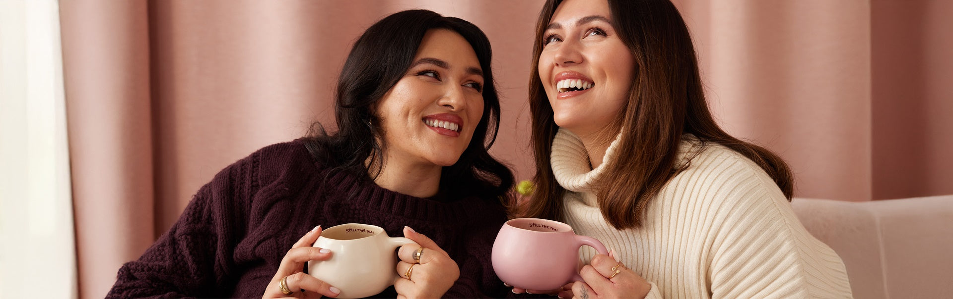 Zwei Frauen sitzen lächelnd auf einem Sofa mit Tassen in der Hand und blicken nach oben.