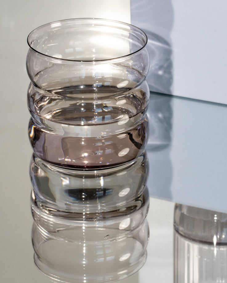 Transparentes, gewelltes Glasgefäß steht auf einem reflektierenden Tisch, umgeben von hellen und dunklen Lichtreflexen.