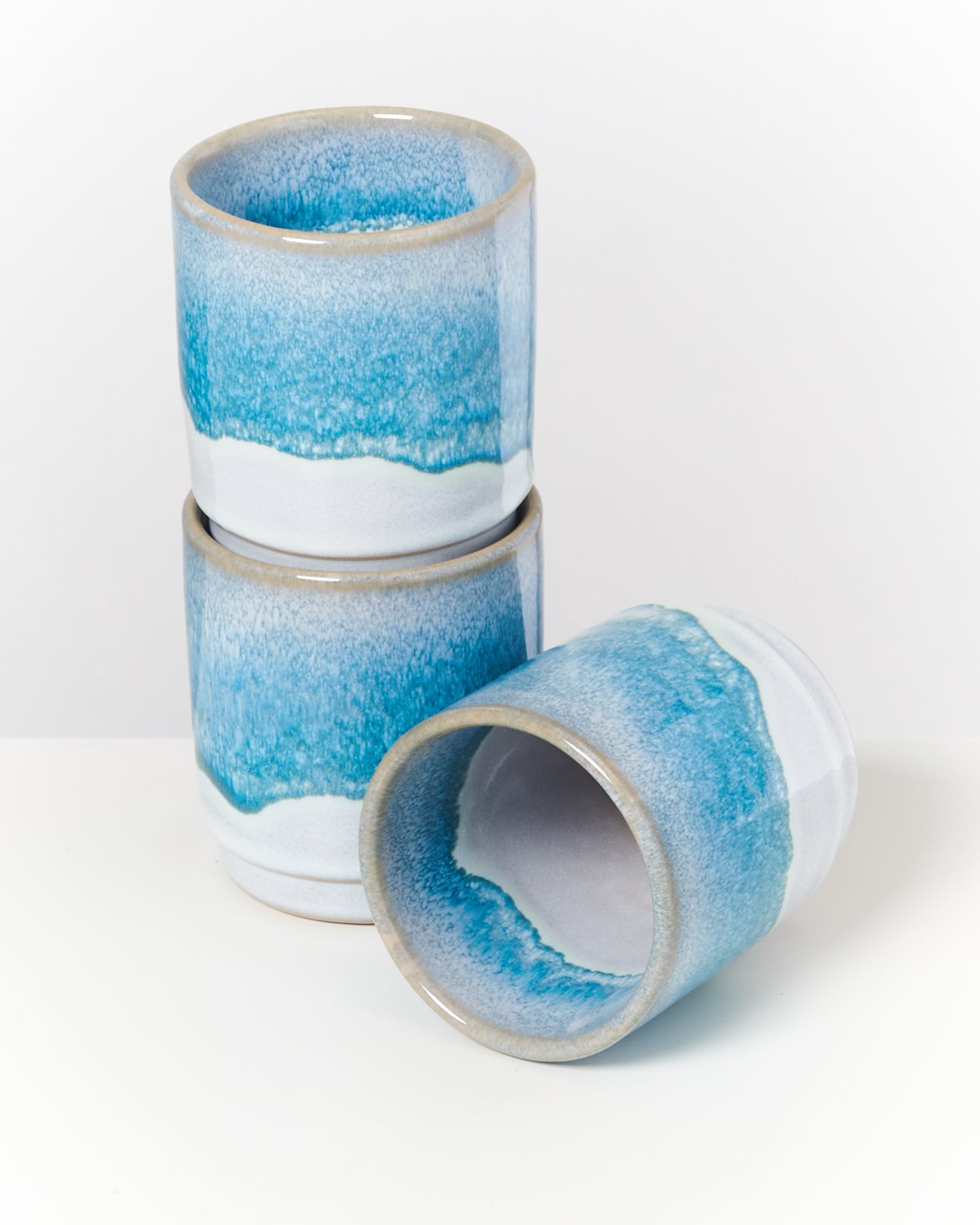 Zwei blaue und weiße Keramiktassen, eine steht, die andere liegt auf der Seite, auf einer weißen Oberfläche.