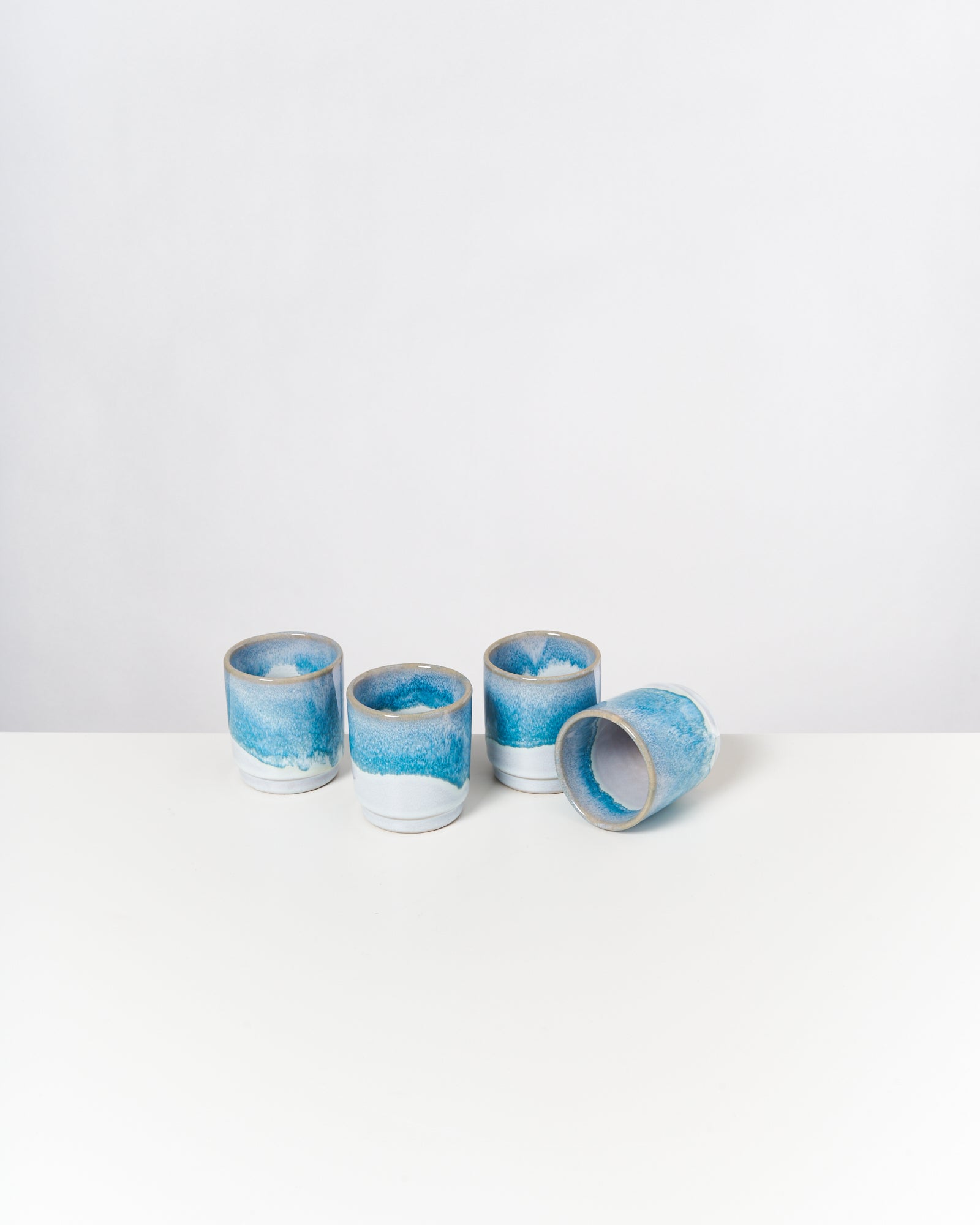 Vier blau-weiße Keramiktassen, eine liegt seitlich, auf einem weißen Tisch vor weißem Hintergrund.