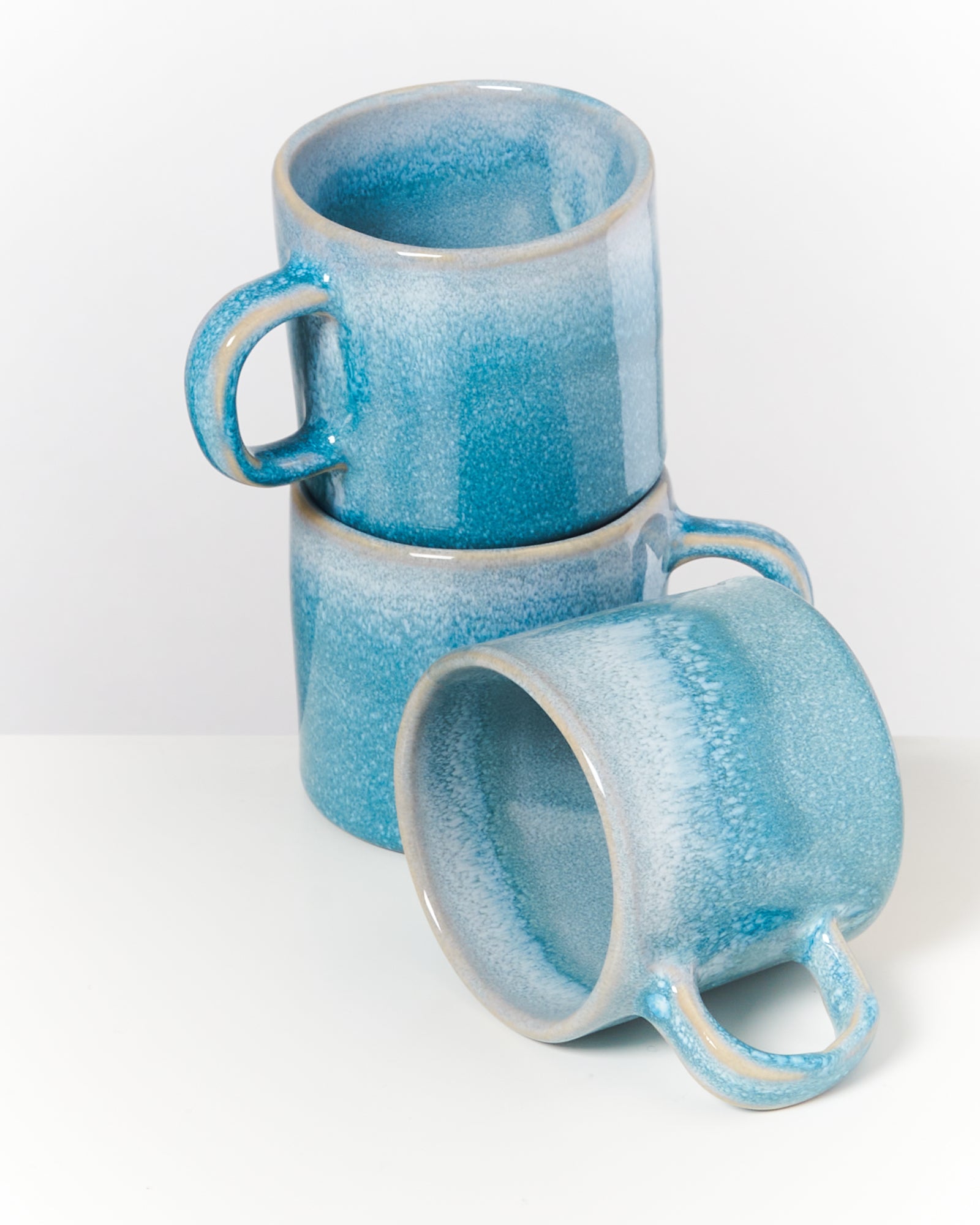 Drei blaue Keramiktassen, zwei gestapelt und eine seitlich liegend, auf einem weißen Hintergrund.