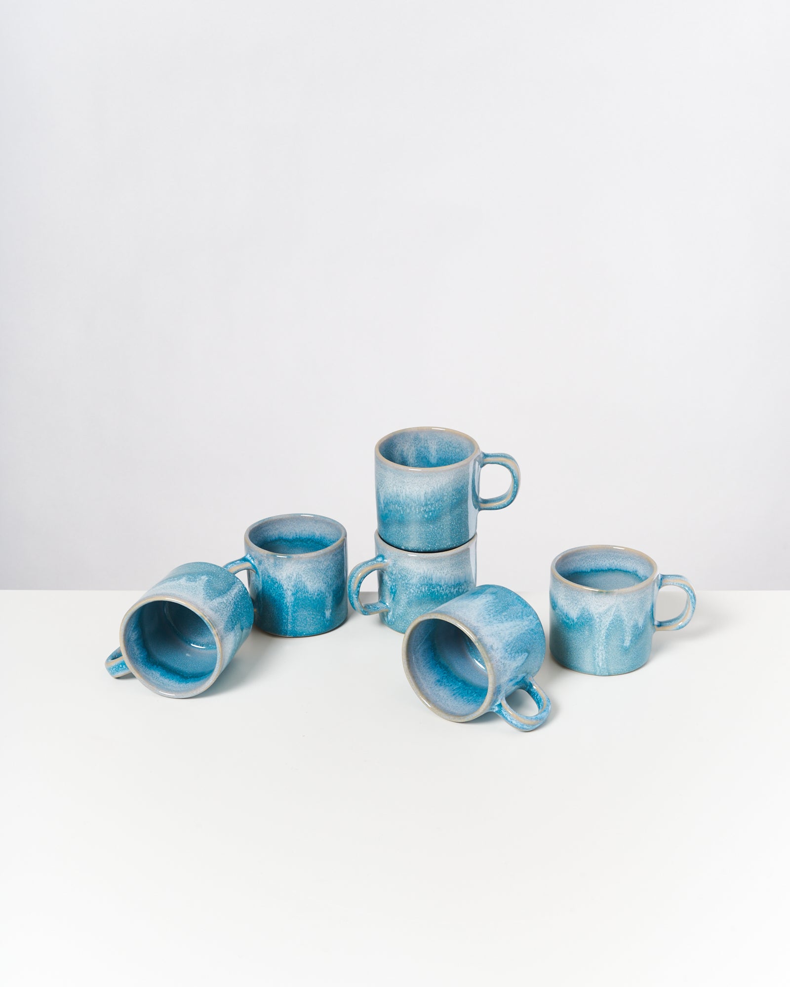Sechs türkisfarbene Tassen mit Henkel sind unregelmäßig auf einem weißen Tisch angeordnet.