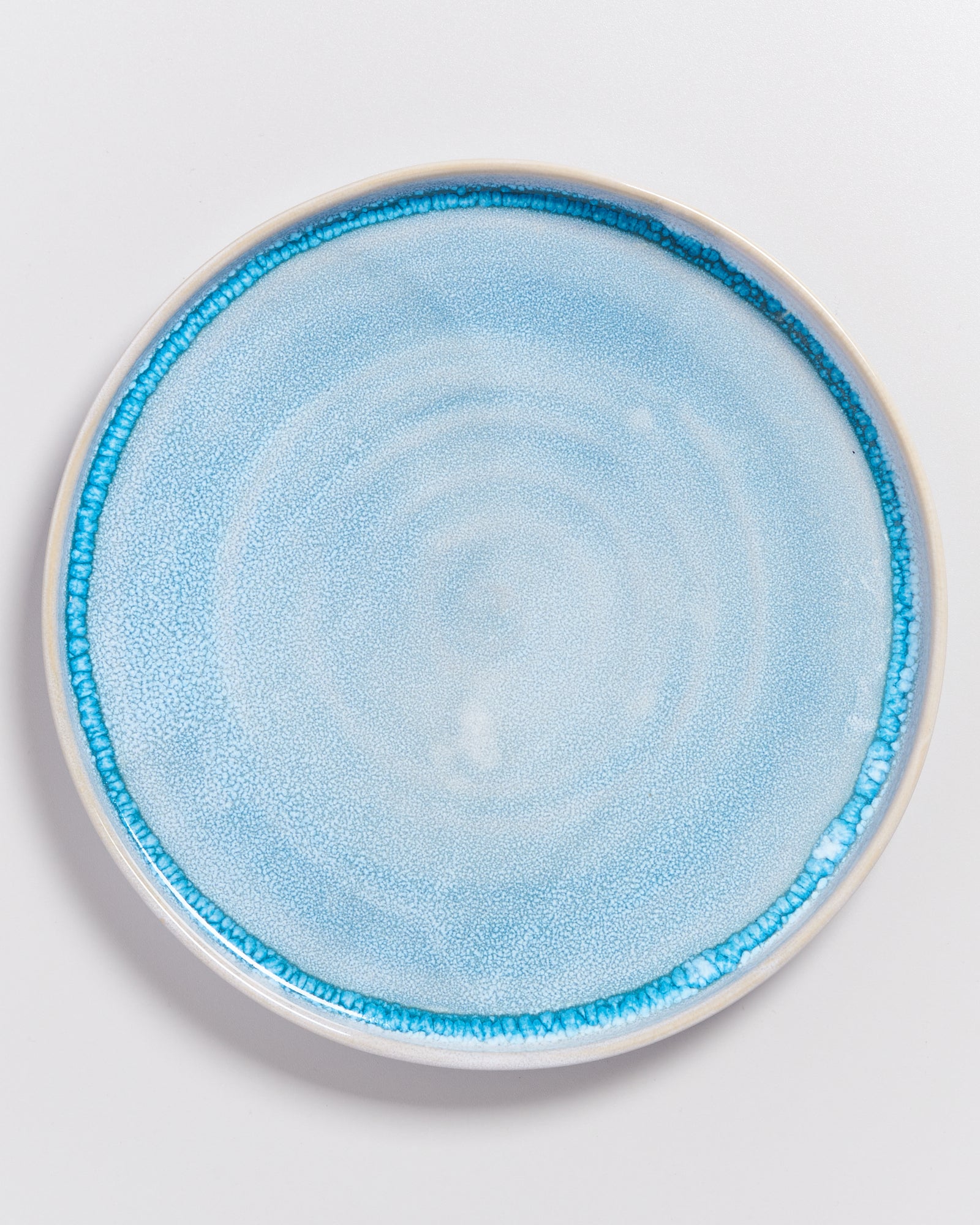 Runder Teller mit hellblauer Glasur und dunklerem Rand auf weißem Hintergrund.