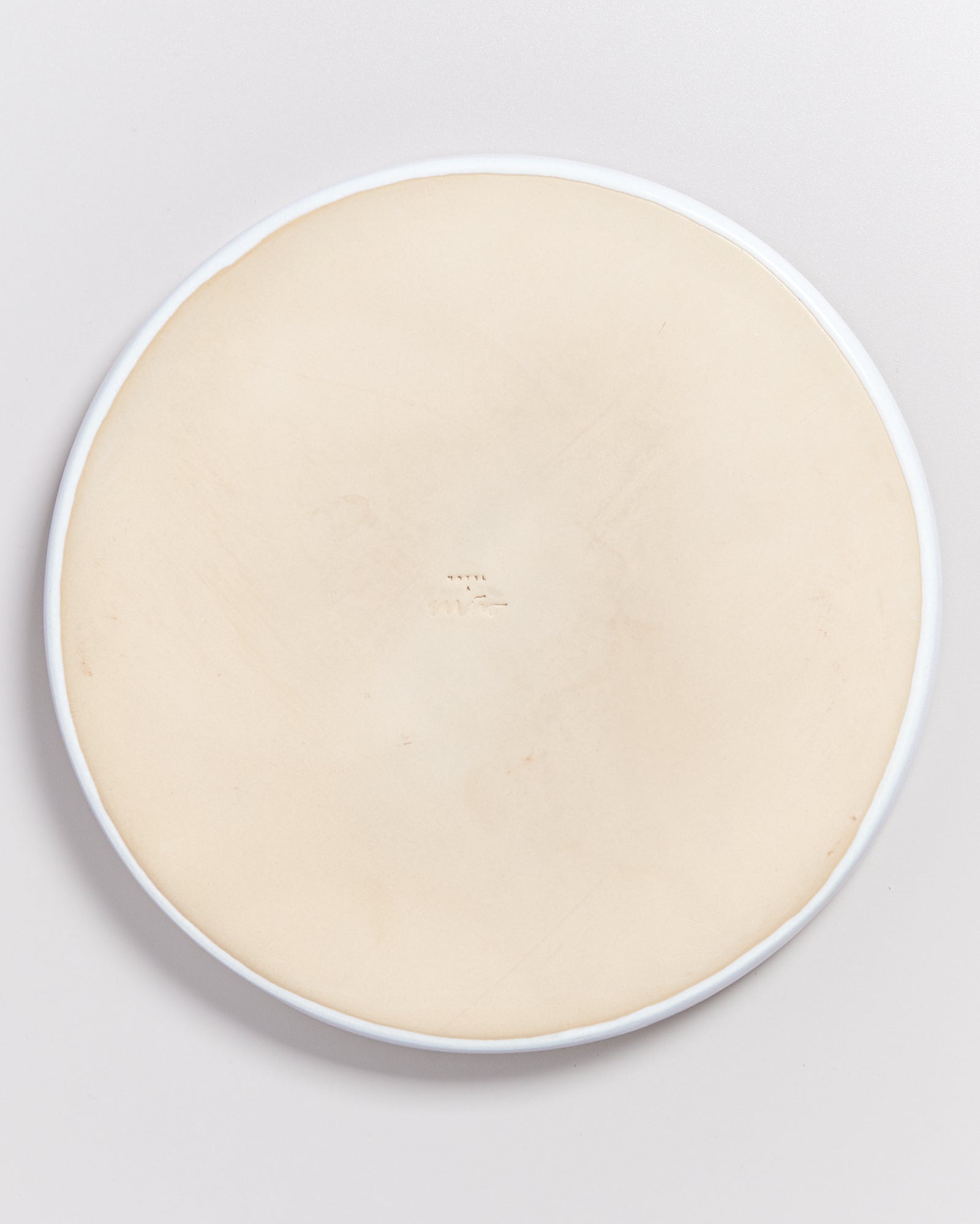 Rückseite eines beigen Keramiktellers mit einer eingeprägten Markierung in der Mitte auf einem hellgrauen Hintergrund.