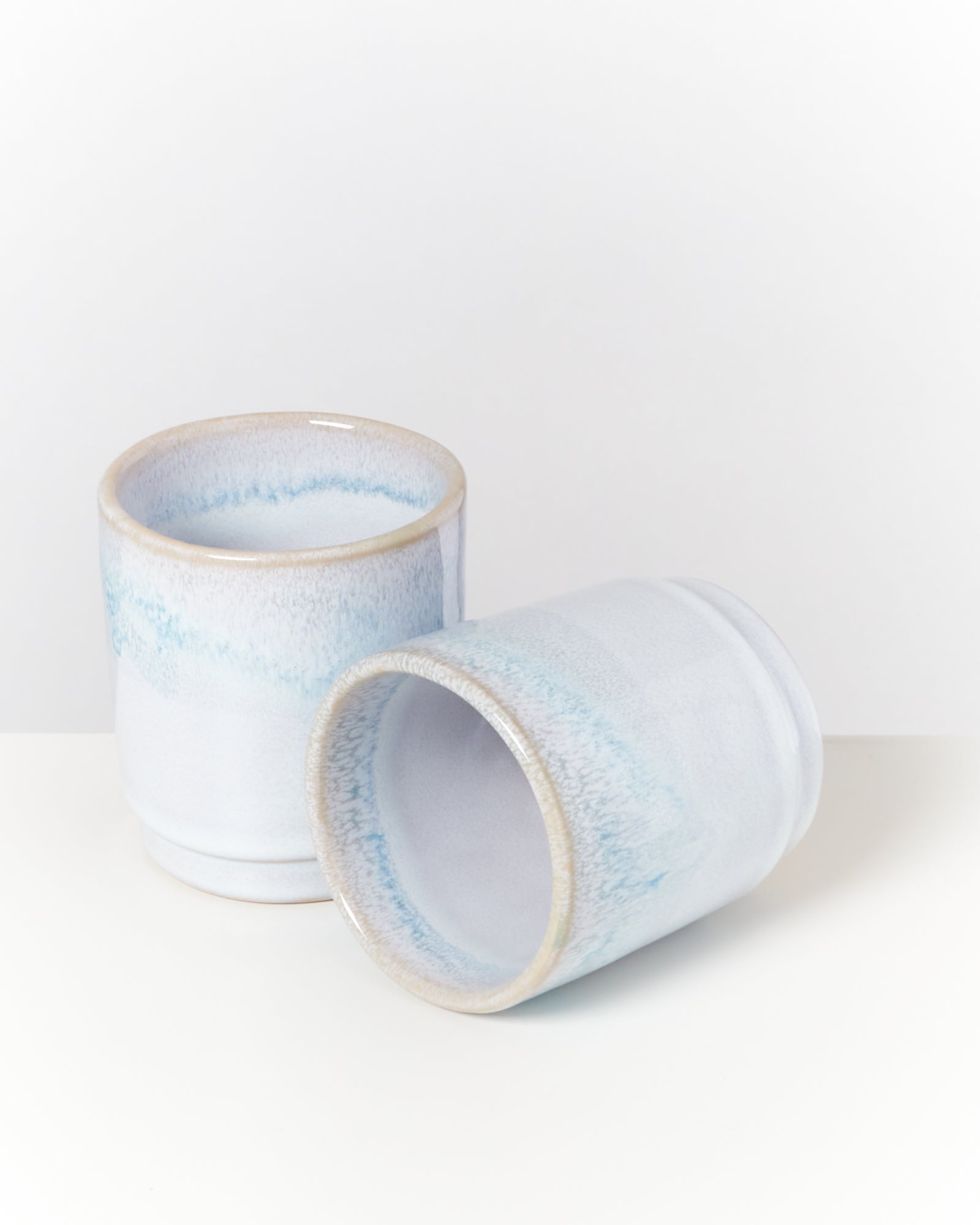 Zwei keramische Becher mit blau-weißer Glasur auf weißem Hintergrund, einer stehend, der andere liegend.