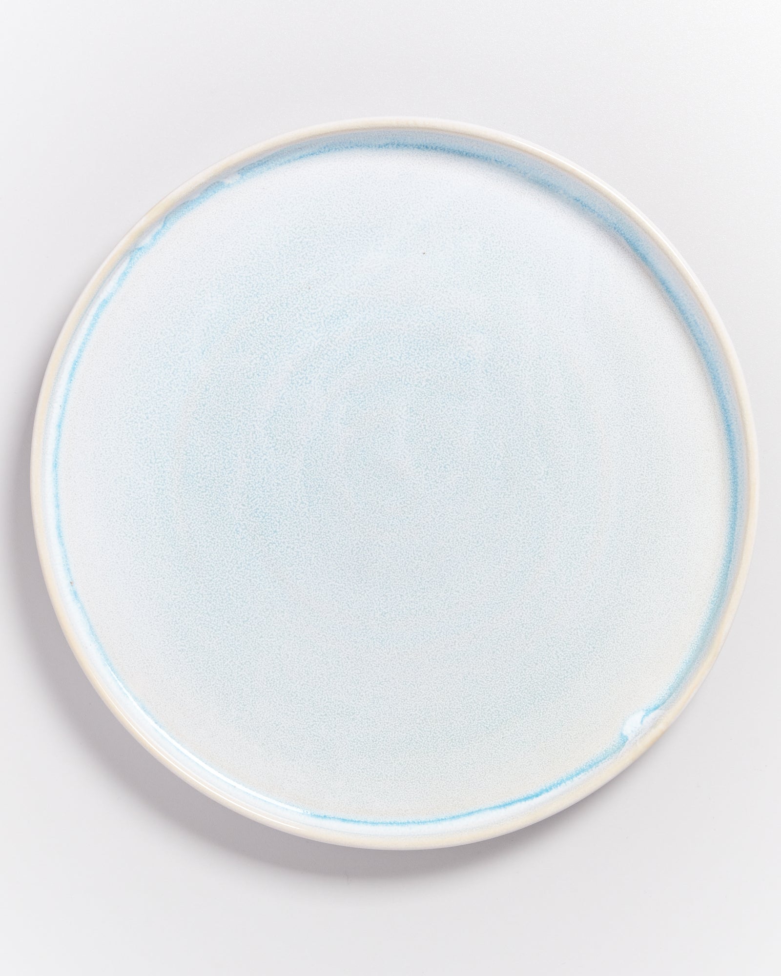 Ein runder, weißer Keramikteller mit einem zarten, blauen Randmuster auf hellem Hintergrund.