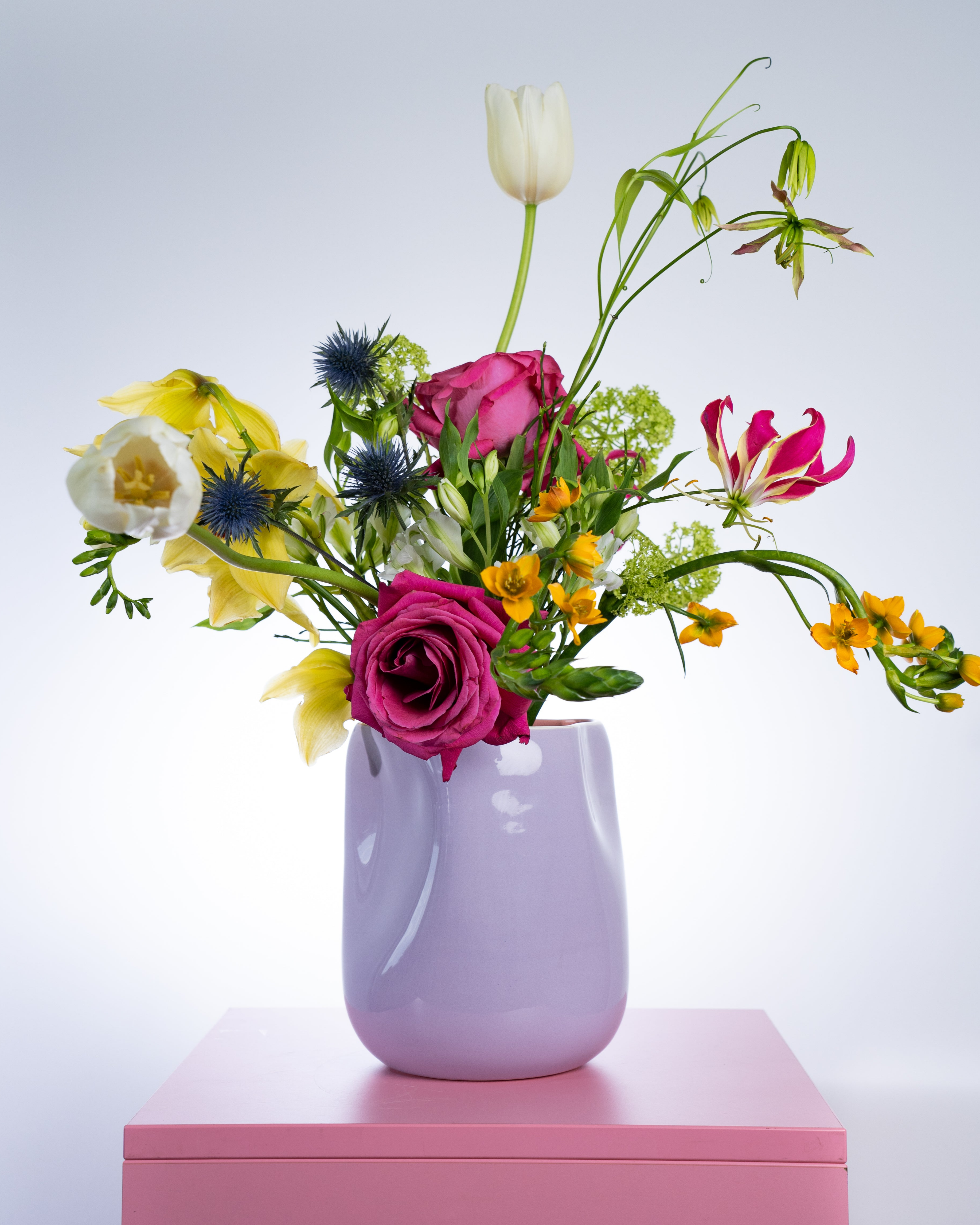 Bunter Blumenstrauß in einer lila Vase auf einem rosa Tisch vor einem neutralen Hintergrund.
