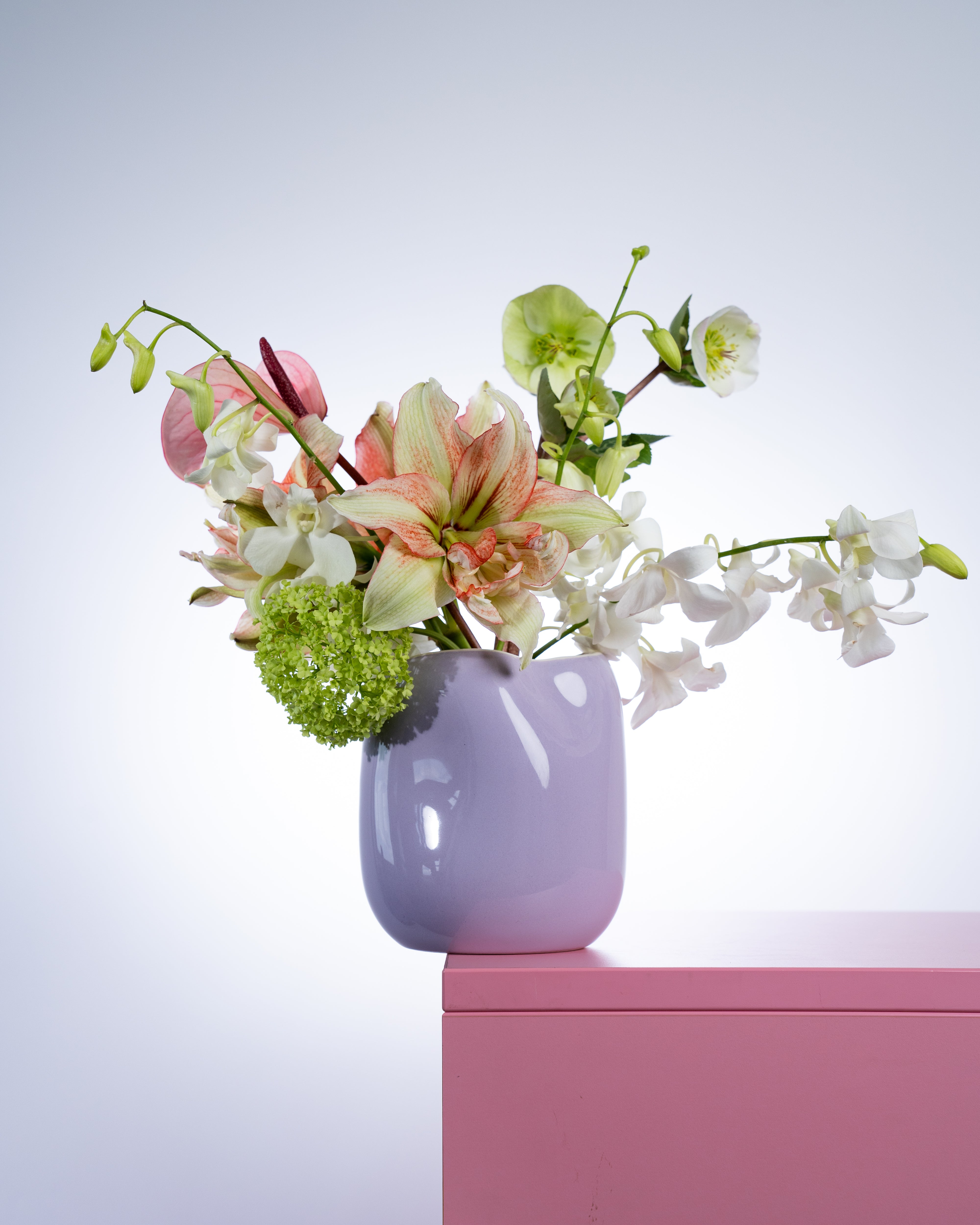 Blumenstrauß in einer lila Vase auf einem pinken Tisch vor neutralem Hintergrund.