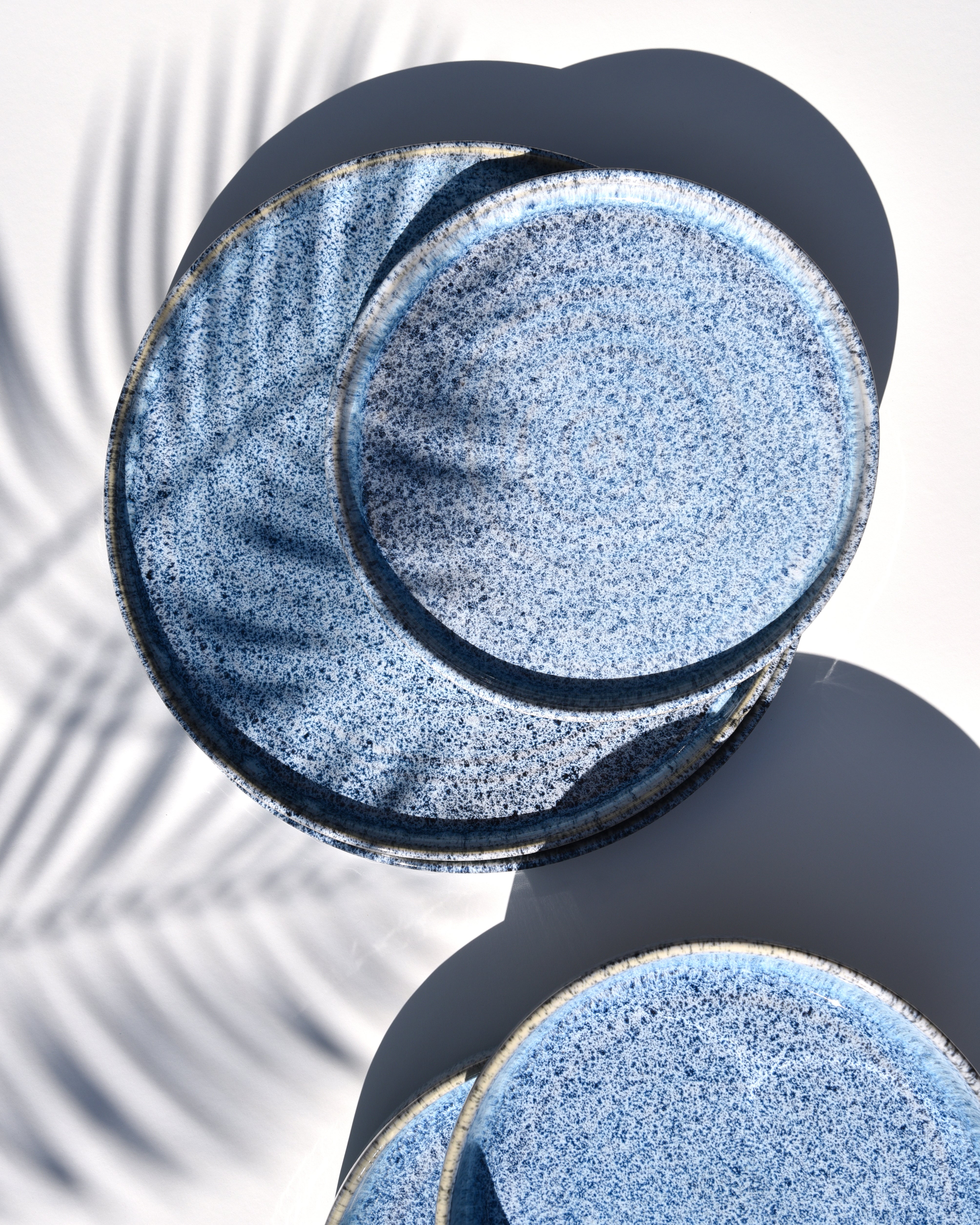 Blaue, gesprenkelte Keramikteller stapeln sich auf weißem Hintergrund mit Schatten einer Palme.