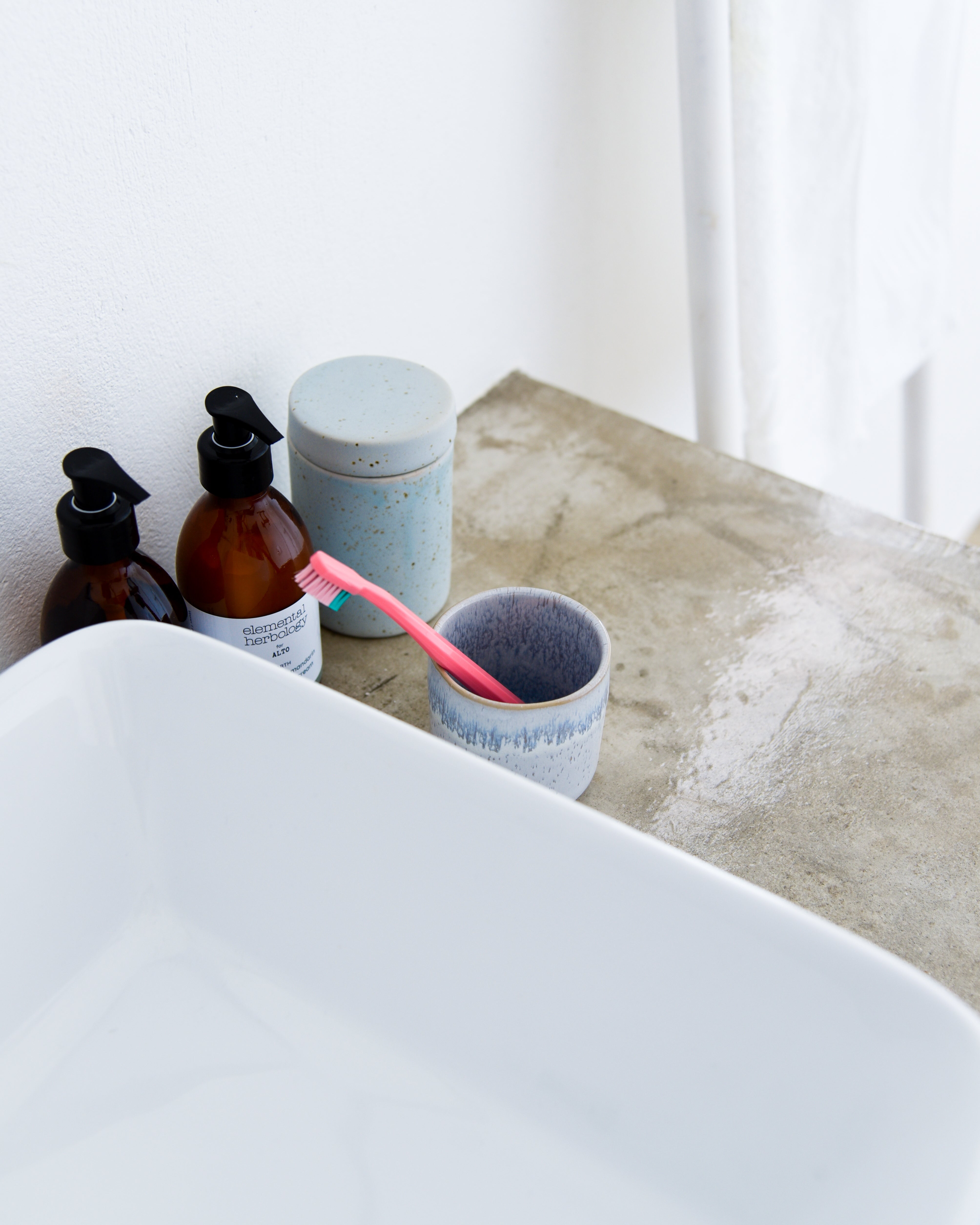 Zahnbürste in einem Becher neben Seifenspendern und einem Keramikbehälter auf einem Badezimmerwaschbecken.