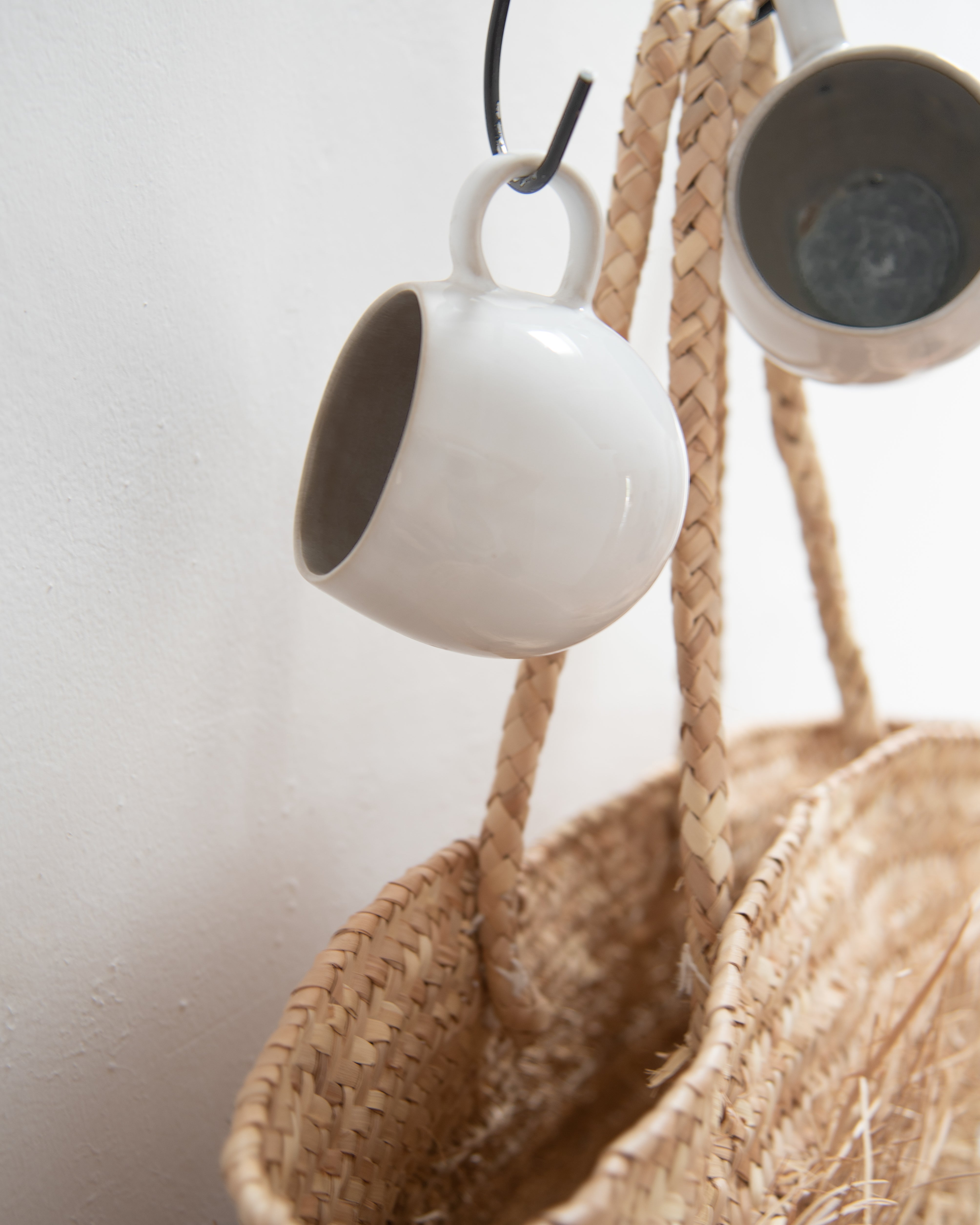 Zwei weiße Tassen hängen an Haken über einem geflochtenen Korb.