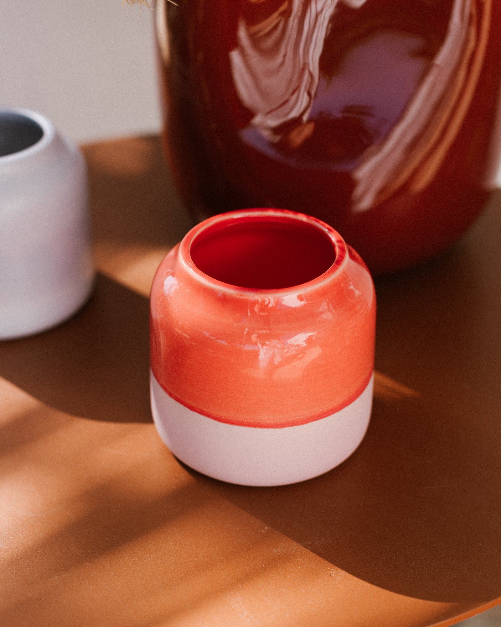 Kleine bunte Vase mit rotem und weißem Design steht auf einem braunen Tisch, umgeben von weiteren Tonkrügen in sanftem Licht.