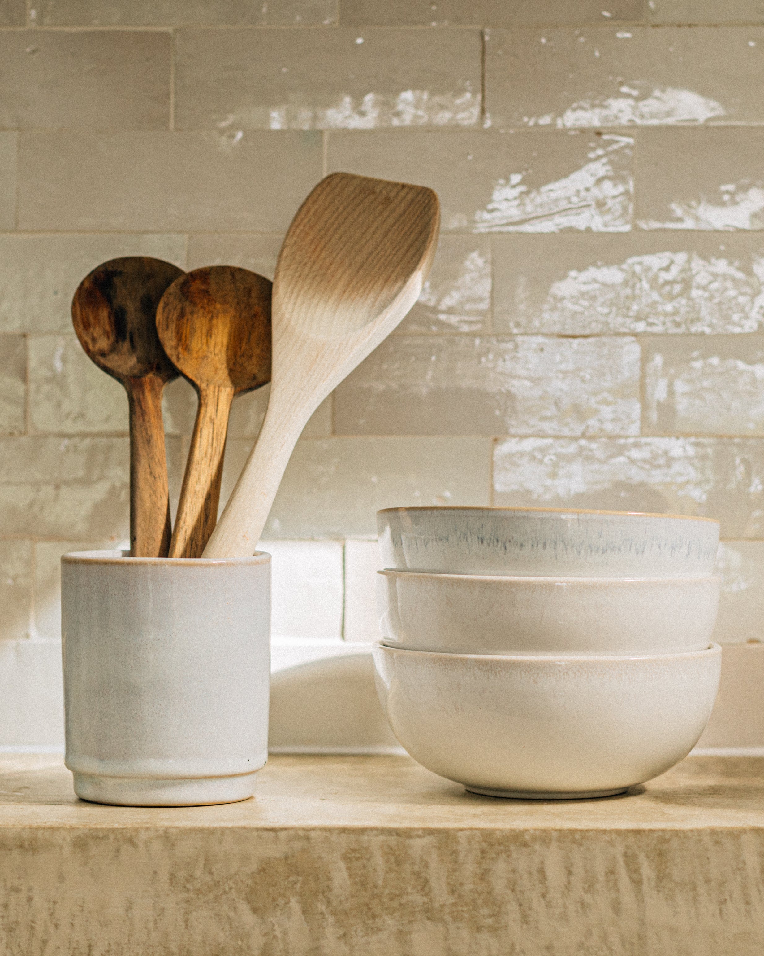 Holzlöffel in einem Keramikbehälter neben drei gestapelten Schalen vor einer Fliesenwand.