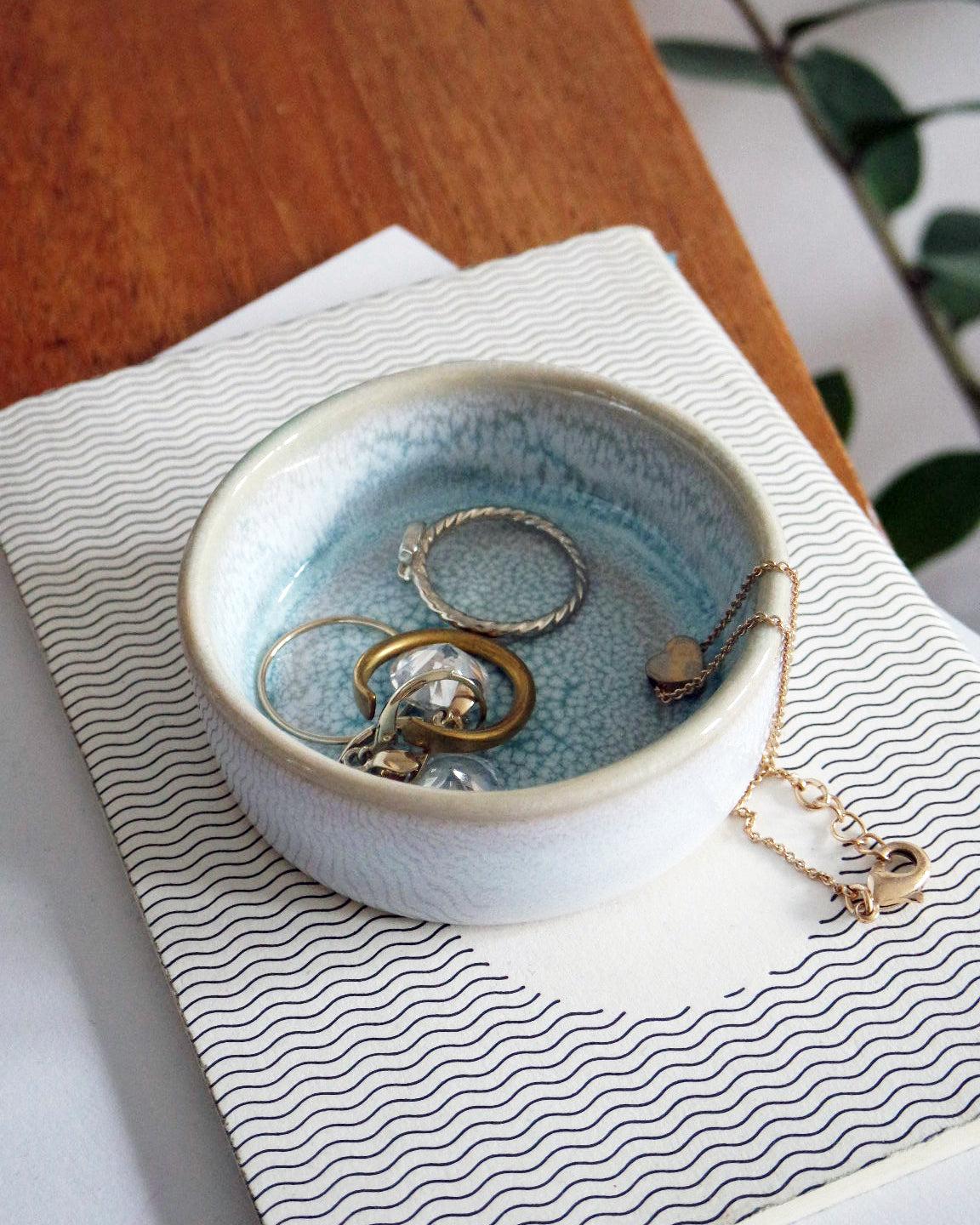 Keramische Schale mit Schmuck, darunter Ringe und Halsketten, steht auf einem Buch mit gewelltem Muster auf einem Holztisch.