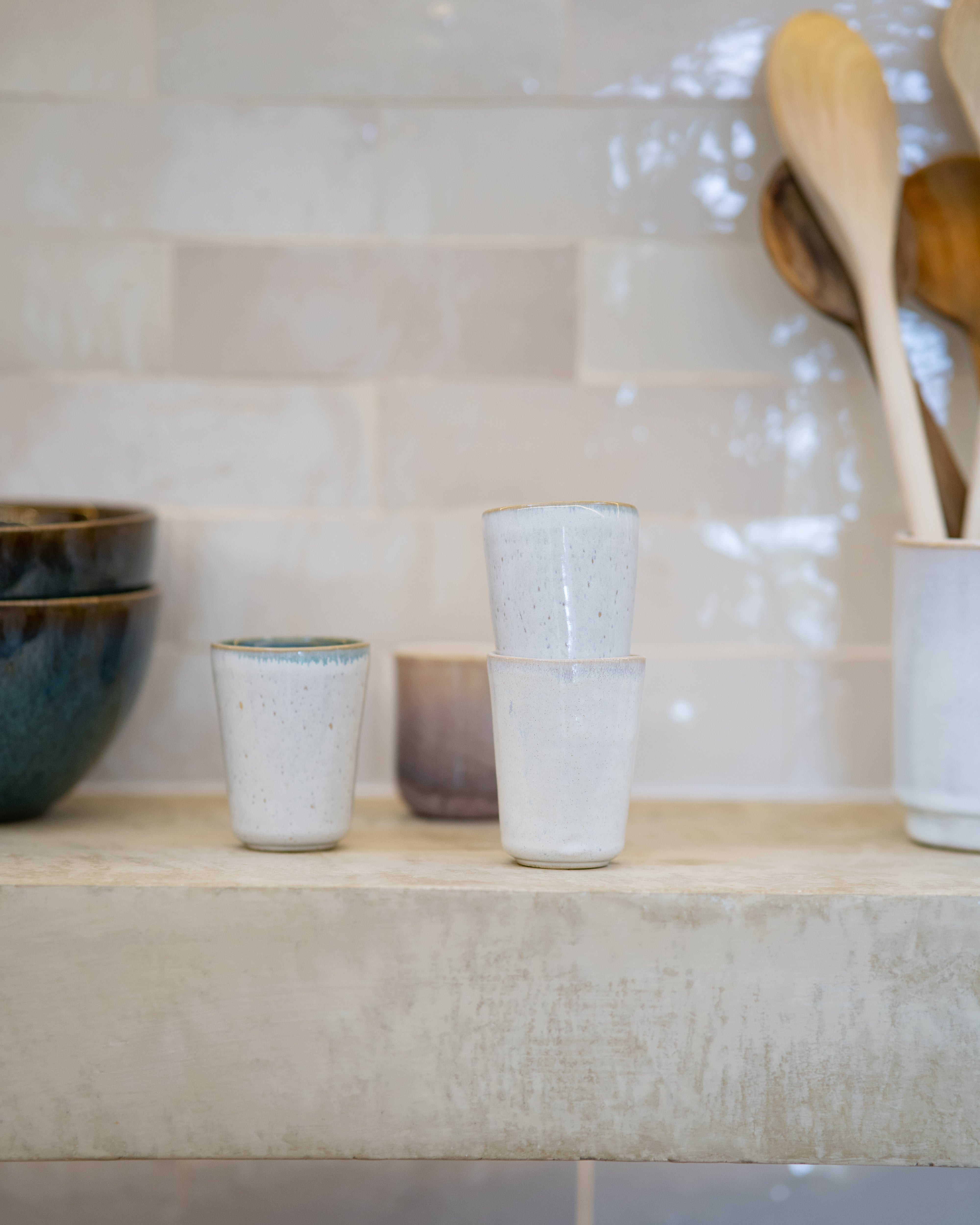 Keramiktassen und Schüsseln auf einem rustikalen Regal vor einer gefliesten Wand, mit hölzernen Küchenutensilien im Hintergrund.