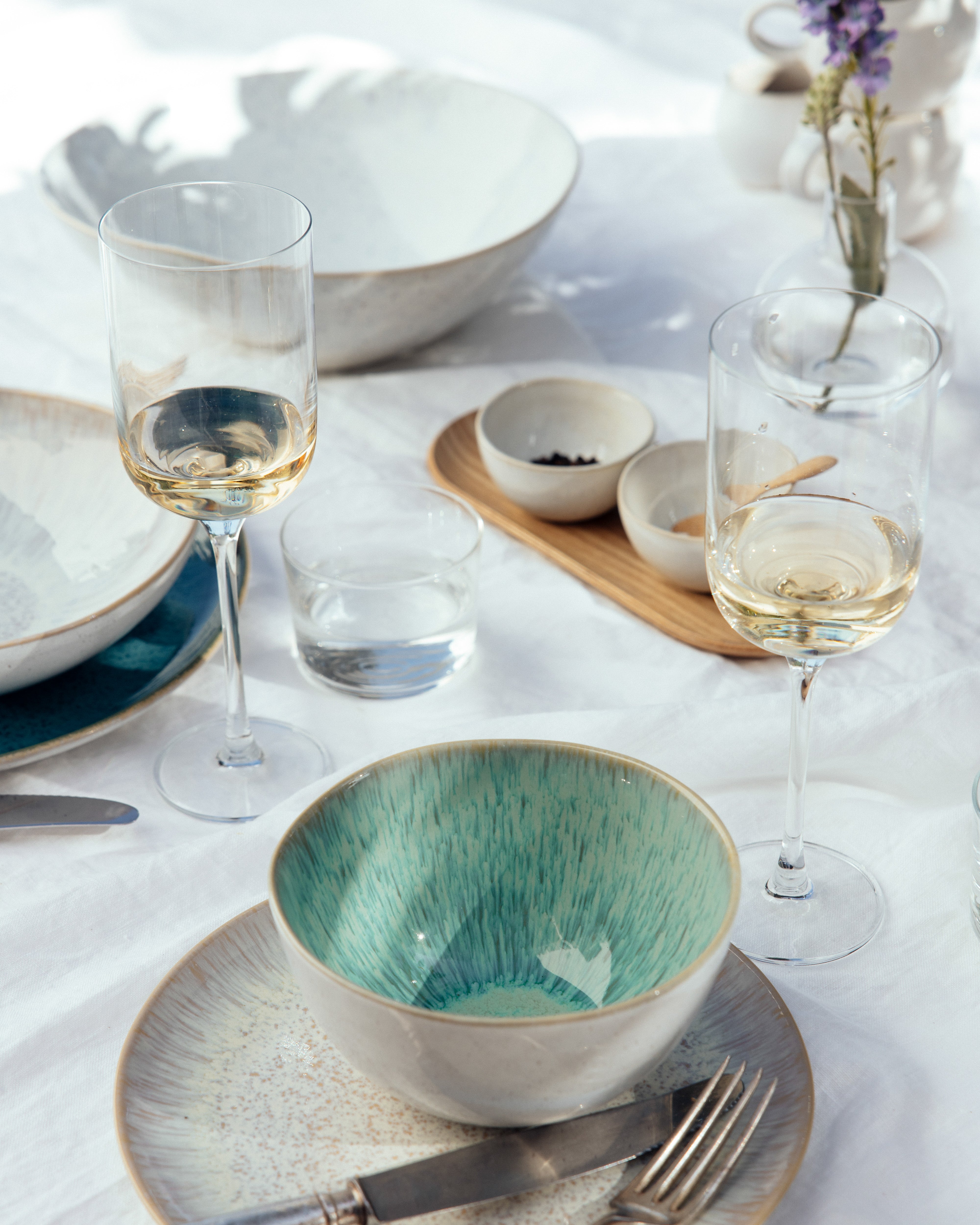 Geschirr auf einem Tisch mit zwei Weingläsern, einer türkisfarbenen Schale, Besteck und kleinen Schalen.