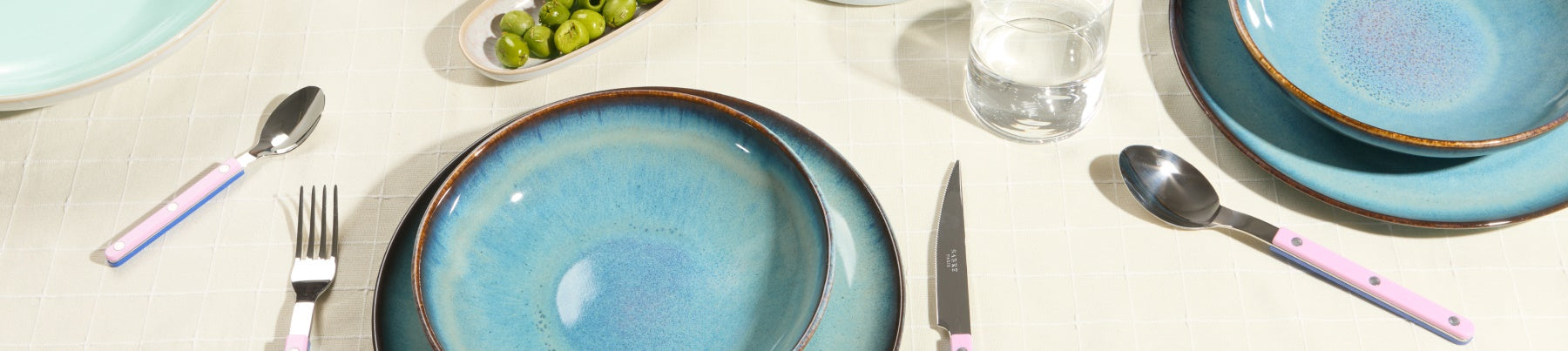 Gedeckter Tisch mit blauen Tellern, rosa Besteck, Glas Wasser und einer Schale Oliven auf beigem Tischdeckchen.