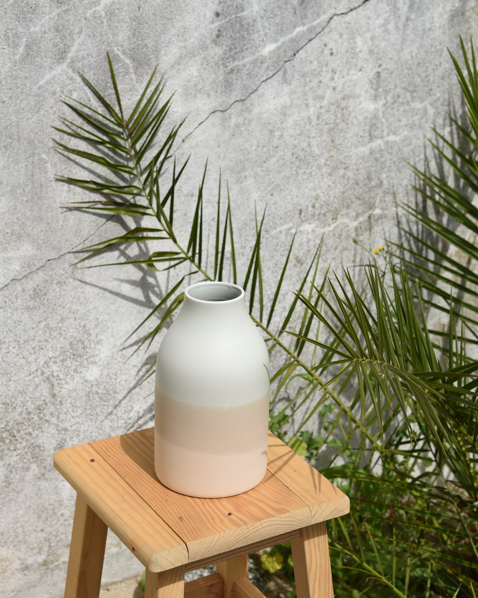 Eine weiß-beige Keramikvase steht auf einem Holzschemel vor einer Betonwand mit grünen Pflanzen.