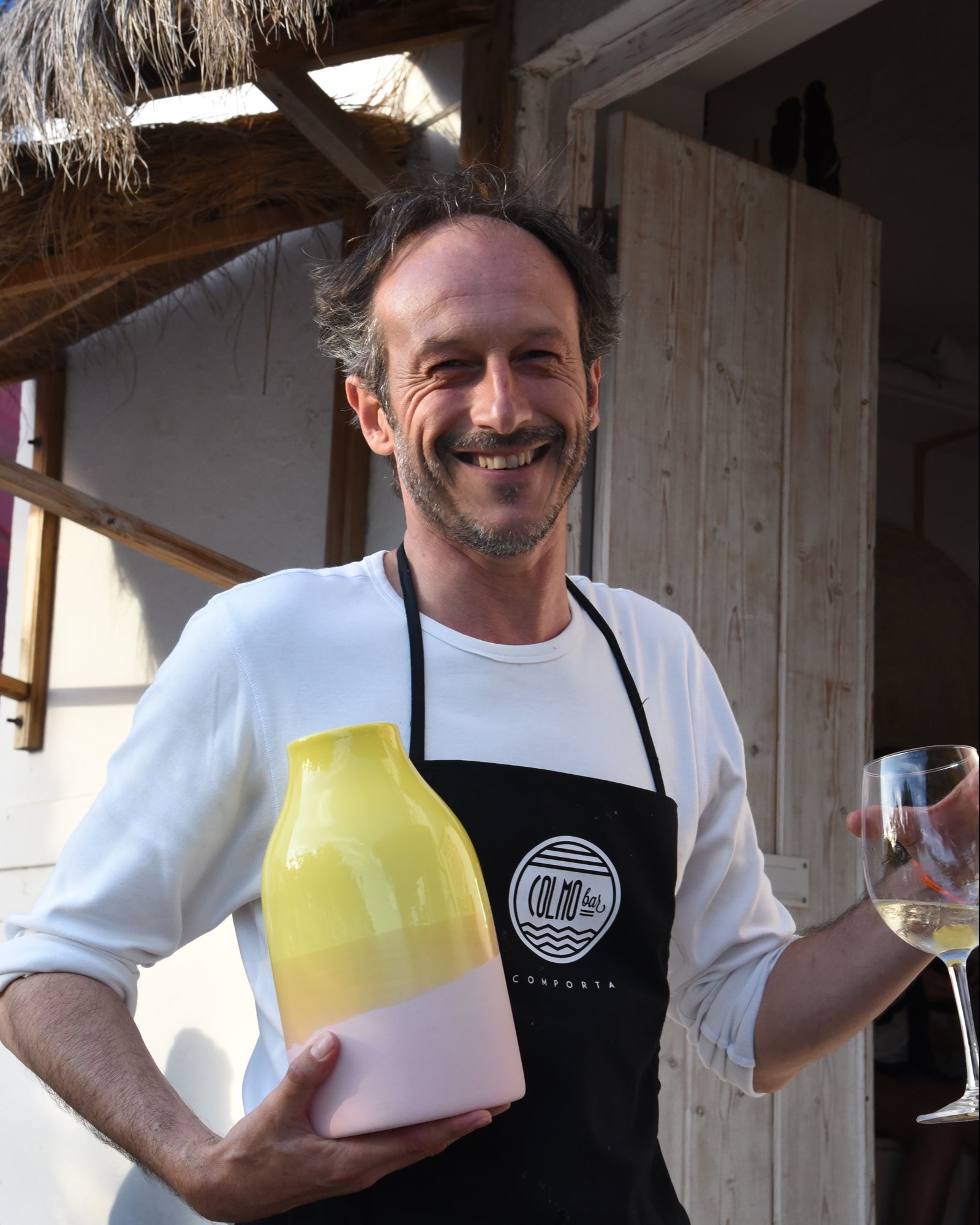Ein lächelnder Mann in Schürze hält eine gelbe Vase und ein Glas Weißwein in der Hand, vor einem Gebäude.