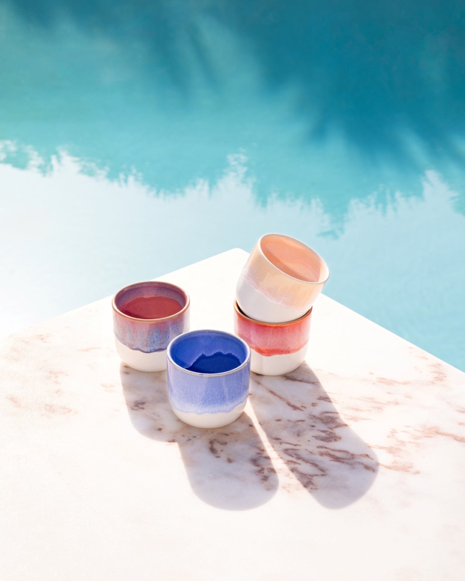 Vier bunte Keramikbecher auf einem Marmortisch in der Sonne, neben einem blauen Pool.