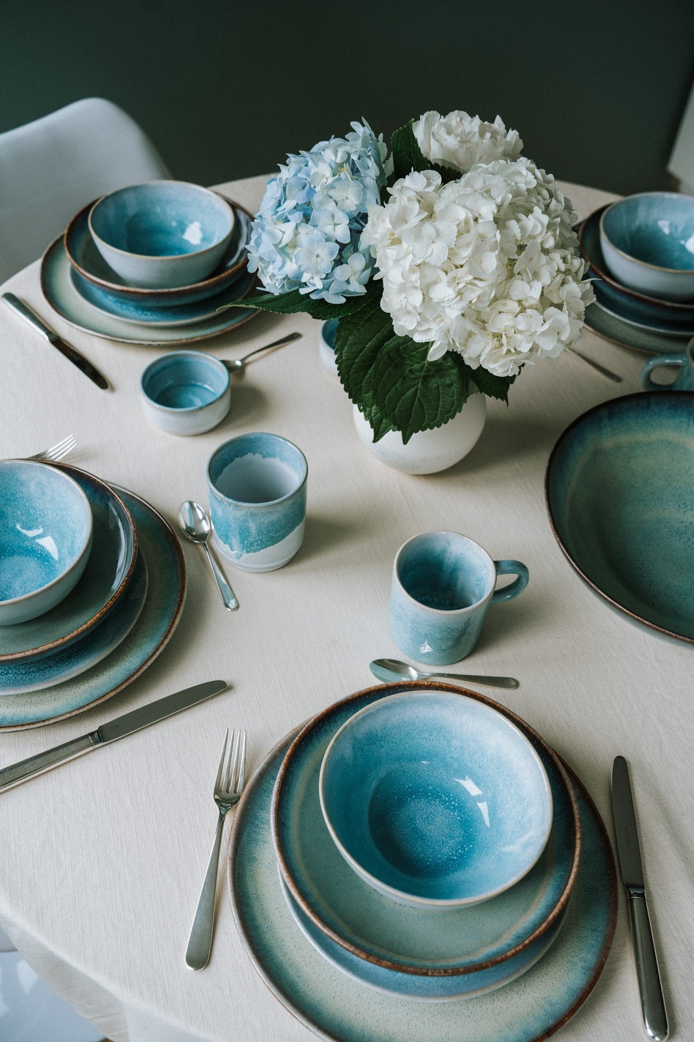 Ein gedeckter Tisch mit blauen Tellern und Tassen, Besteck und einer Vase mit blauen und weißen Hortensien.
