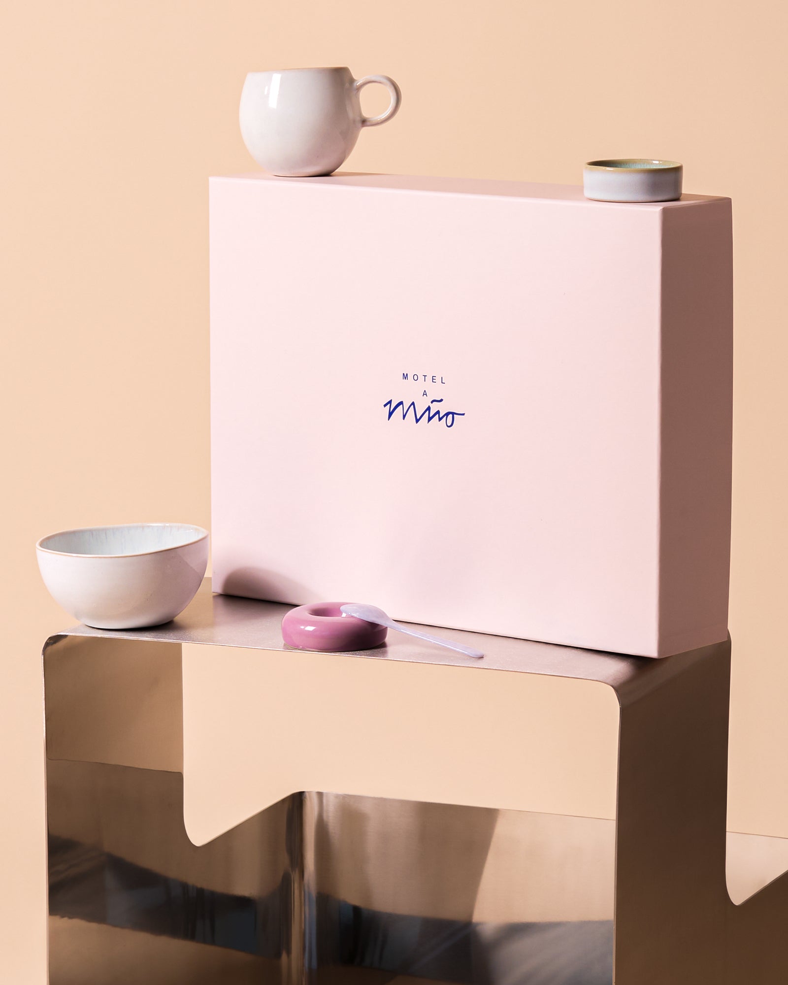 Rosa Schachtel mit der Aufschrift "Motel a Miio" auf einem Tisch mit Geschirr; minimalistisches Design.