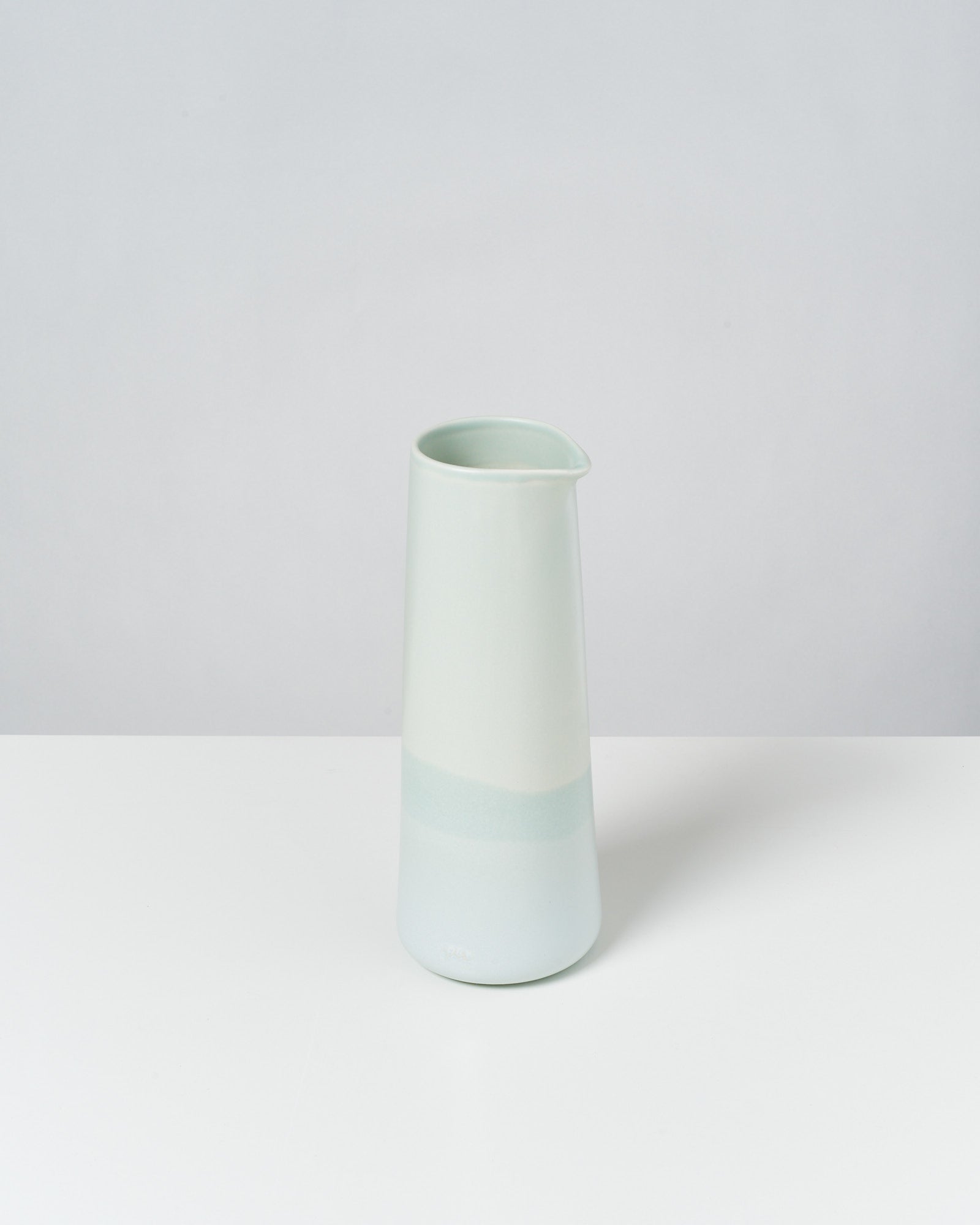 Heller, pastellfarbener Krug aus Keramik mit schlichter, konischer Form auf einer weißen Oberfläche vor neutralem Hintergrund.