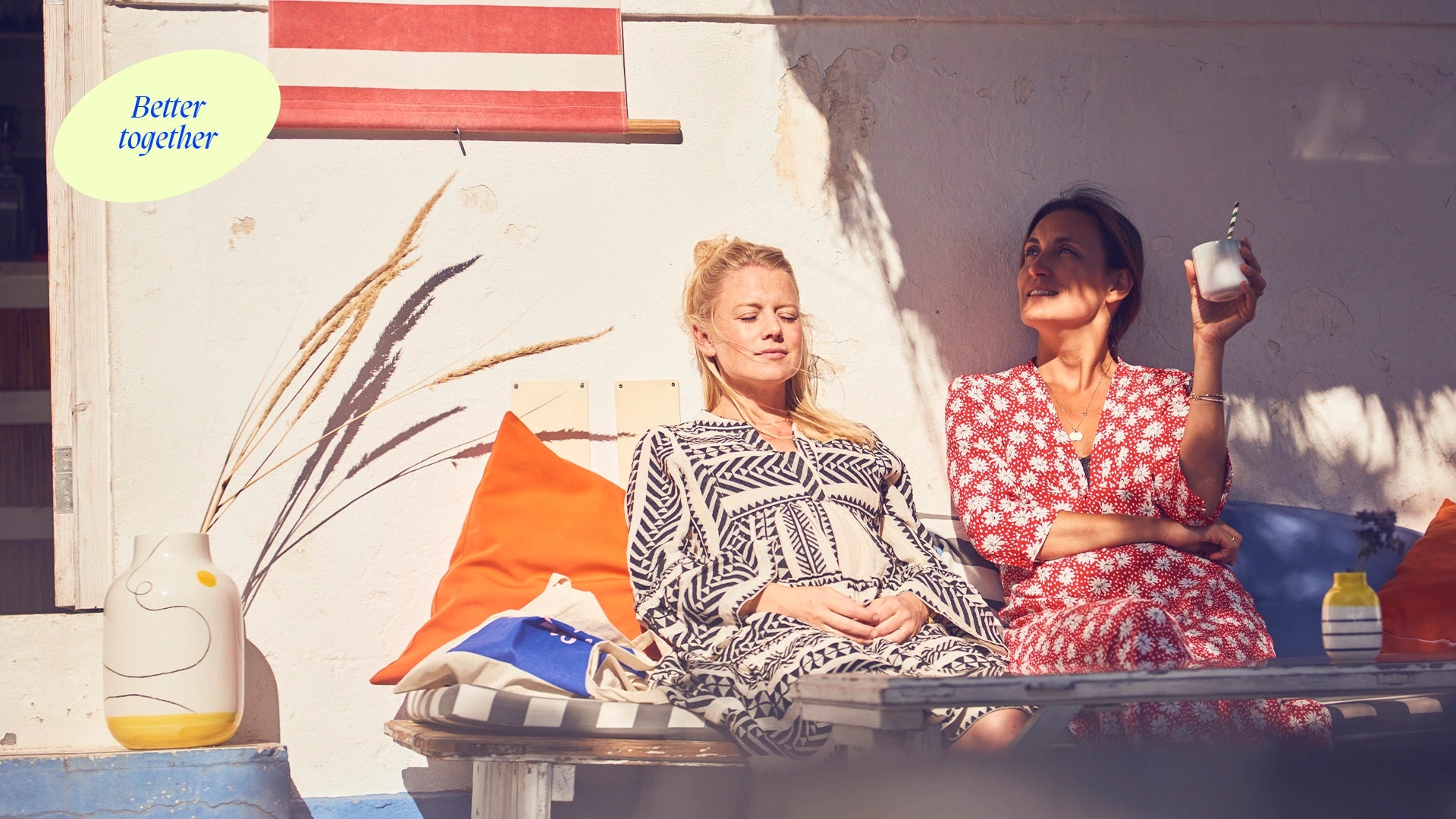 Zwei Frauen entspannen in der Sonne auf einer Bank, umgeben von Kissen und Blumenvasen.