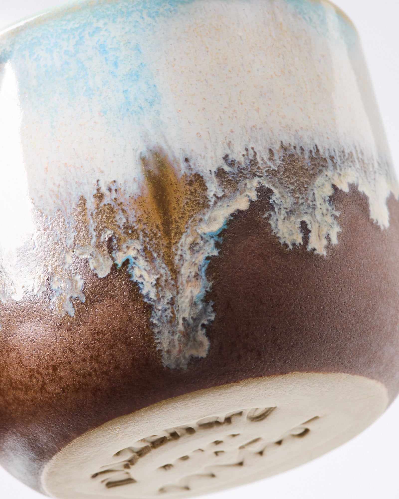 Nahaufnahme einer braunen Keramikvase mit blau-weißen glasigen Verzierungen und einem geprägten Muster am Boden.