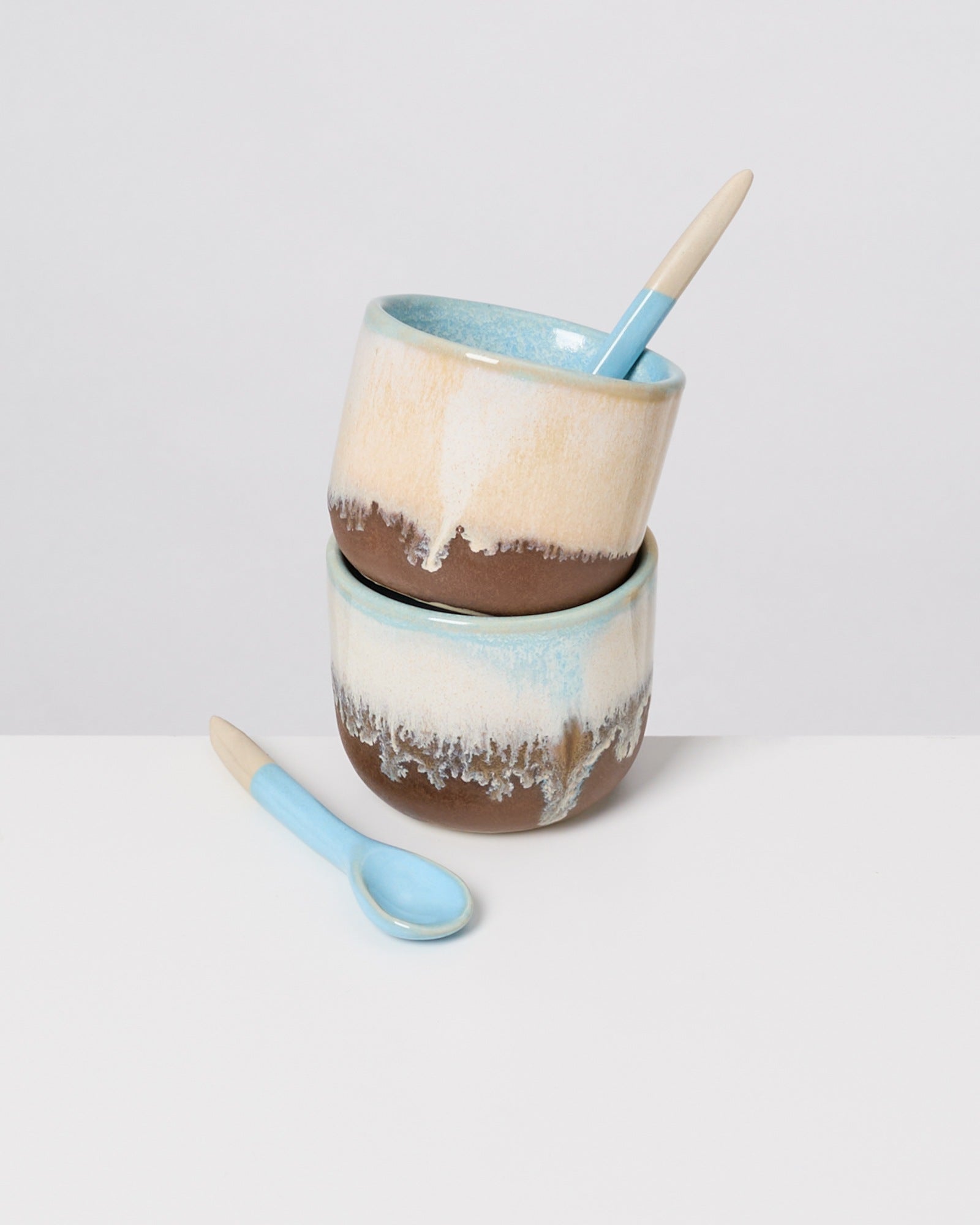 Zwei gestapelte, handgemachte Keramikschalen mit einem blauen Löffel in der oberen Schale auf weißem Hintergrund.
