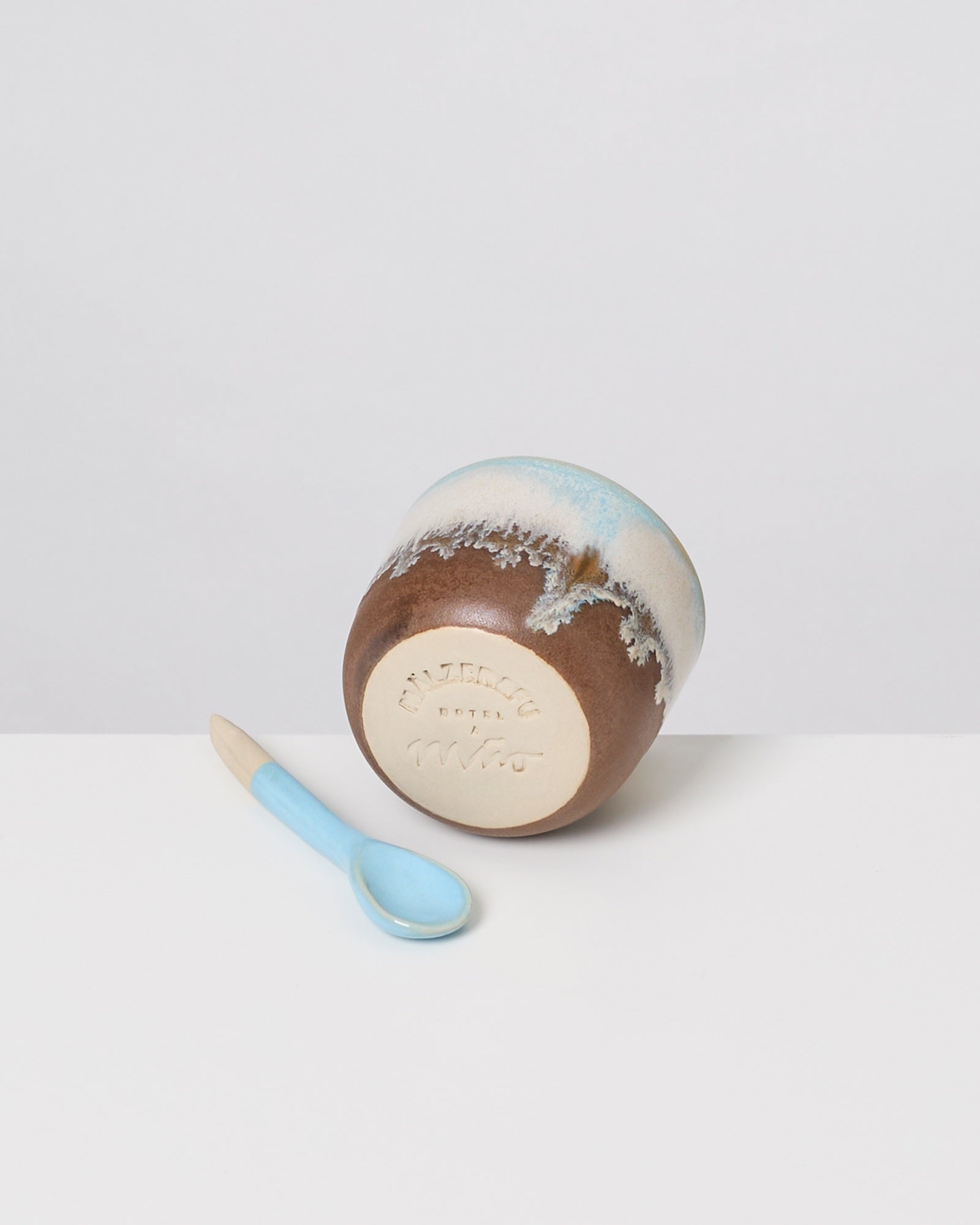 Blau-weißer Löffel neben einer umgedrehten braun-weißen Schale mit strukturierter Oberfläche auf weißem Hintergrund.