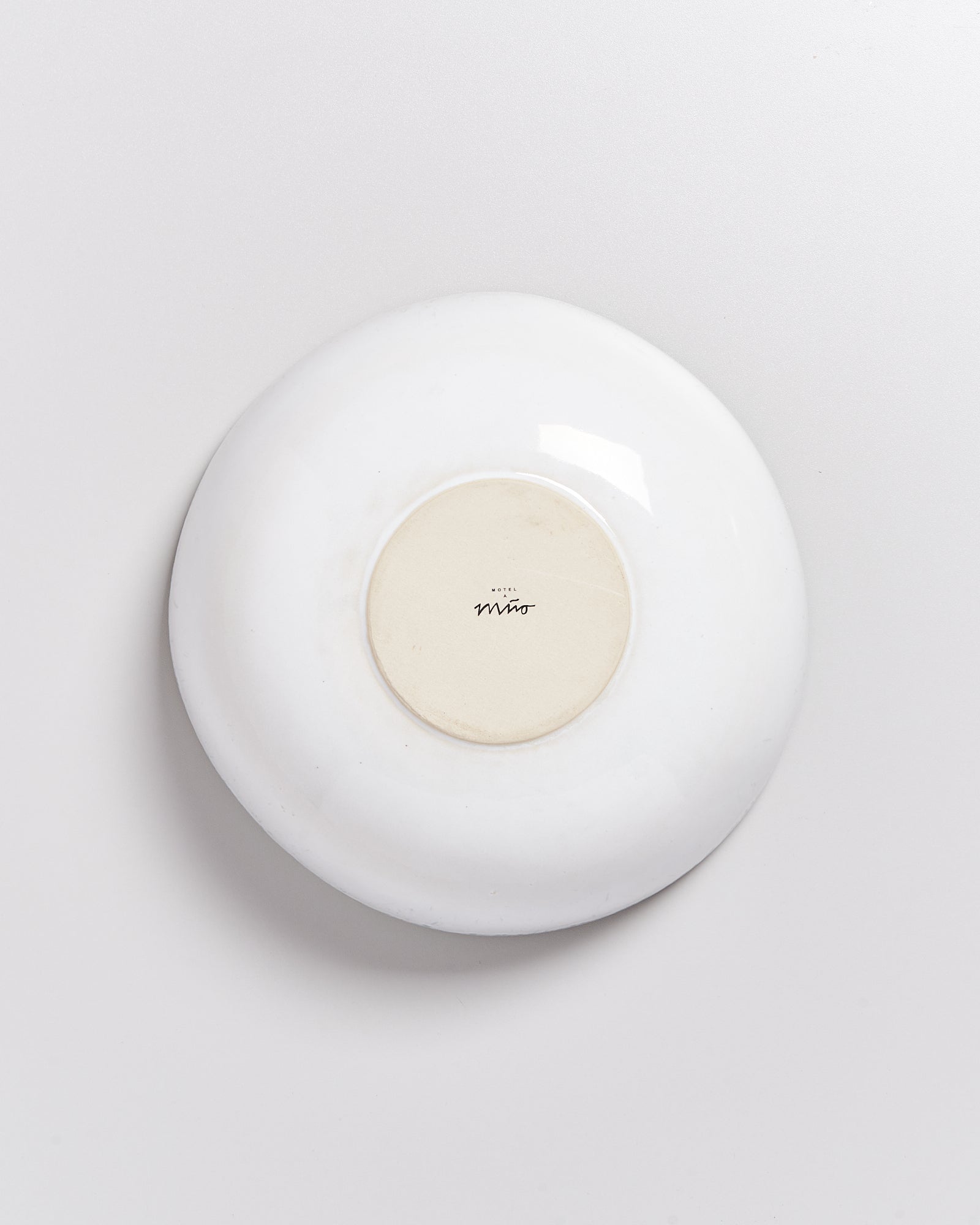 Eine weiße, umgedrehte Keramikschale mit einem runden, beigen Zentrum und einem kleinen schwarzen Schriftzug darauf.