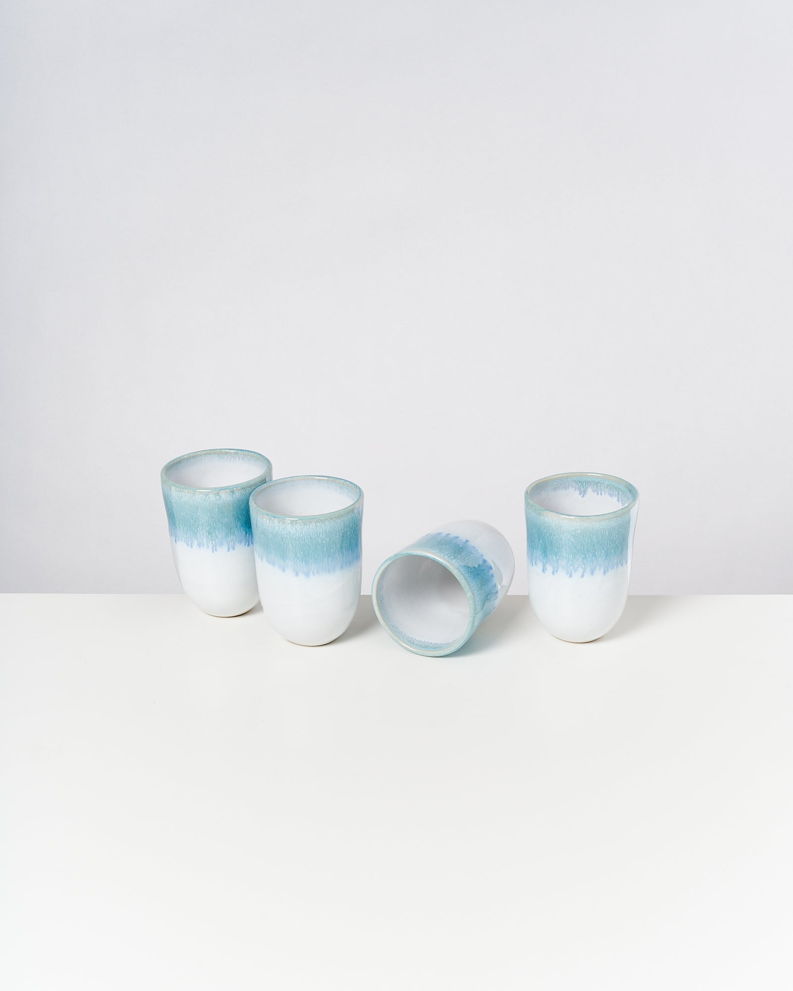 Vier weiß-blaue Keramiktassen, eine liegt seitlich, auf einer weißen Oberfläche vor neutralem Hintergrund.