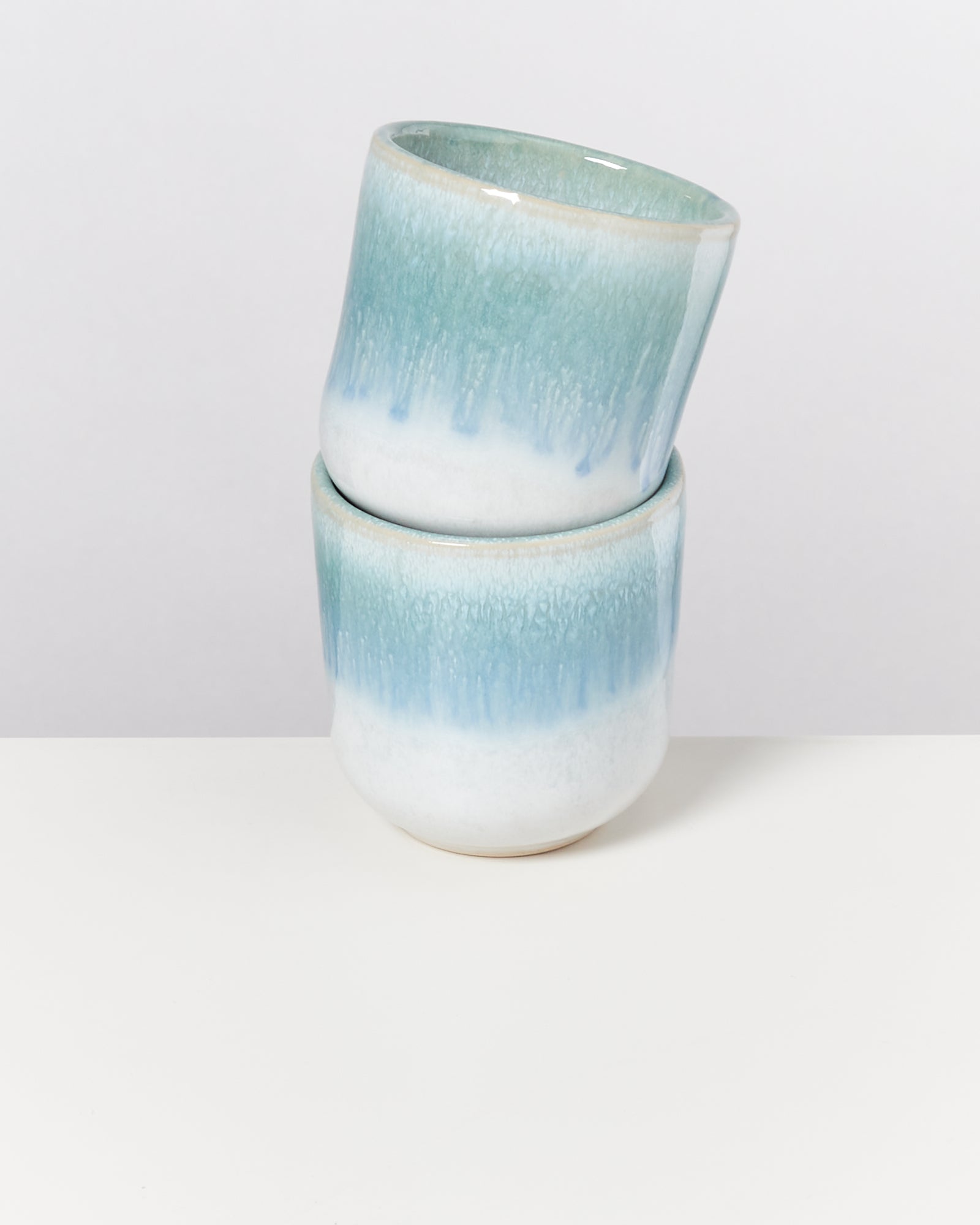 Zwei türkis-weiße, ineinander gestapelte Keramikbecher stehen auf einem weißen Untergrund.