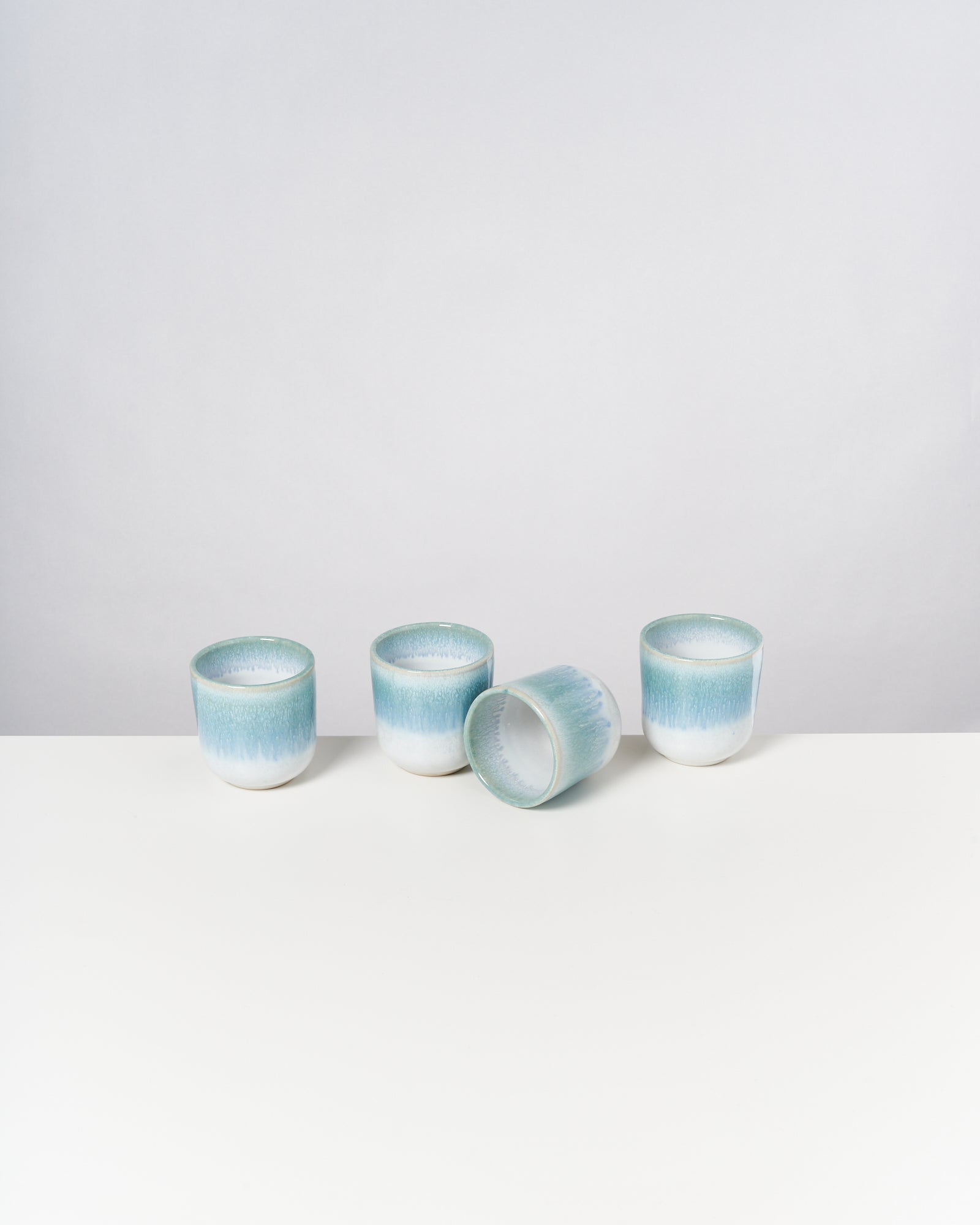 Vier pastellfarbene Keramikbecher stehen auf einem weißen Tisch, einer liegt auf der Seite.