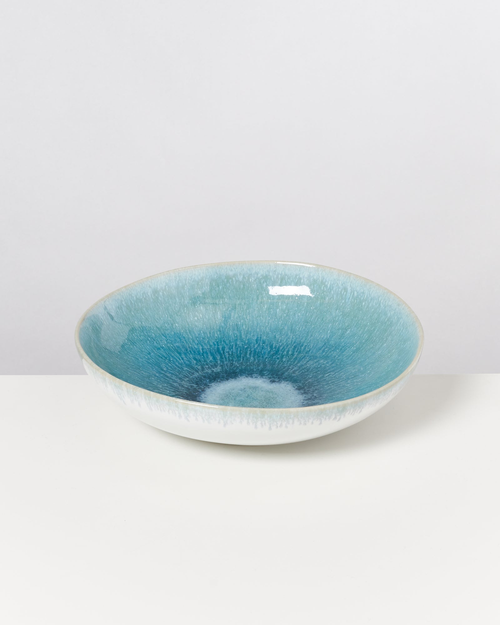Eine flache Schale mit blau-grüner Keramikglasur steht auf einem weißen Untergrund.