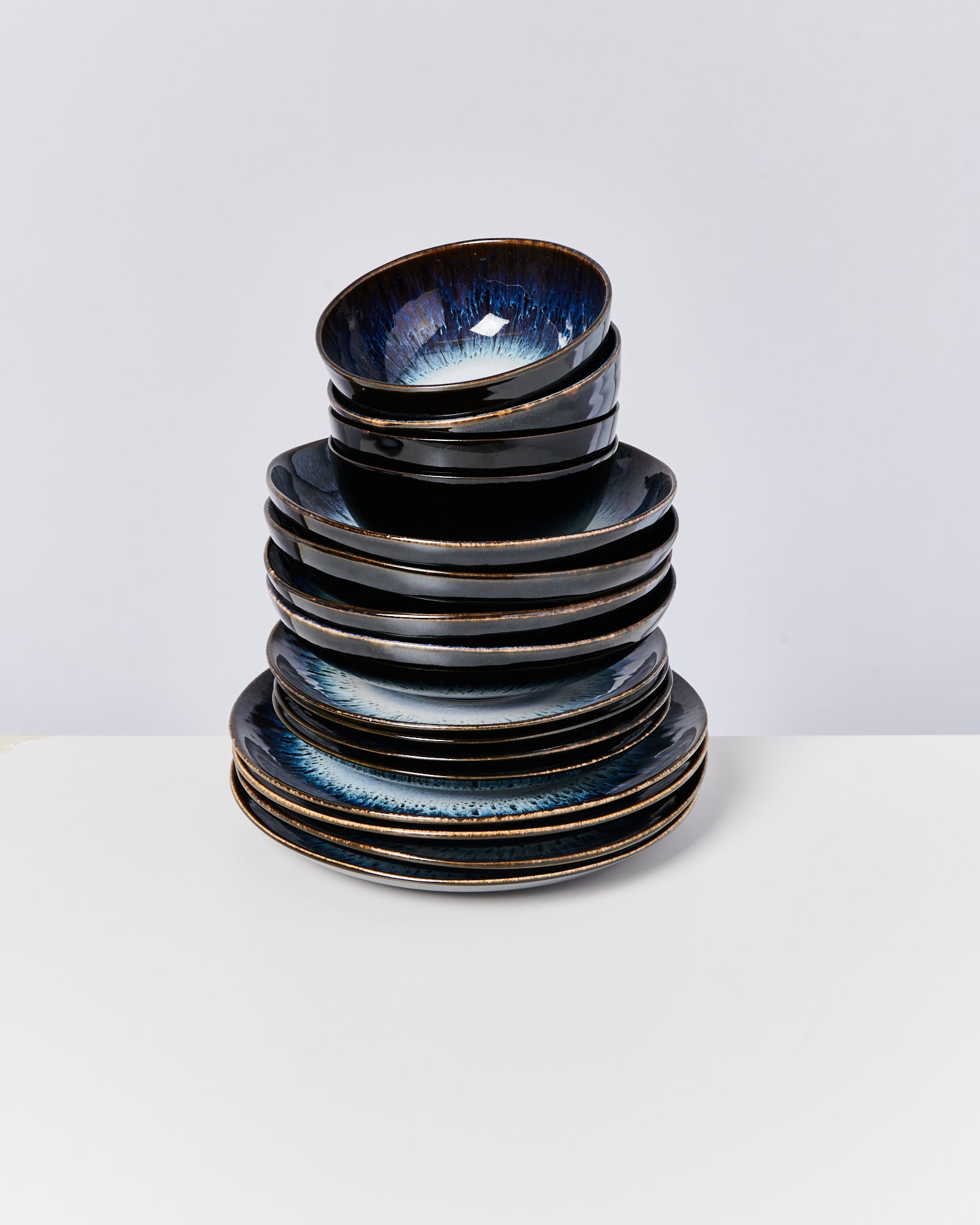 Ein Stapel aus schwarzen Tellern und Schalen mit blauer Innenglasur, auf einem weißen Tisch vor neutralem Hintergrund.