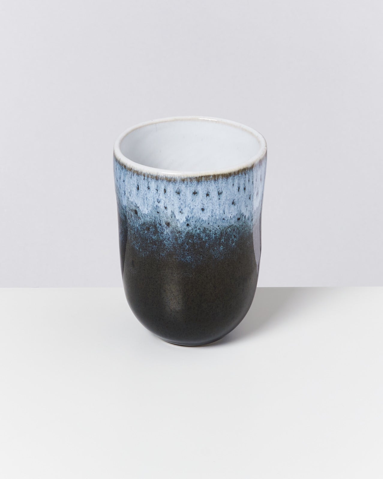 Keramikbecher mit blau-schwarzem Verlaufsdesign auf heller Oberfläche vor einem neutralen Hintergrund.