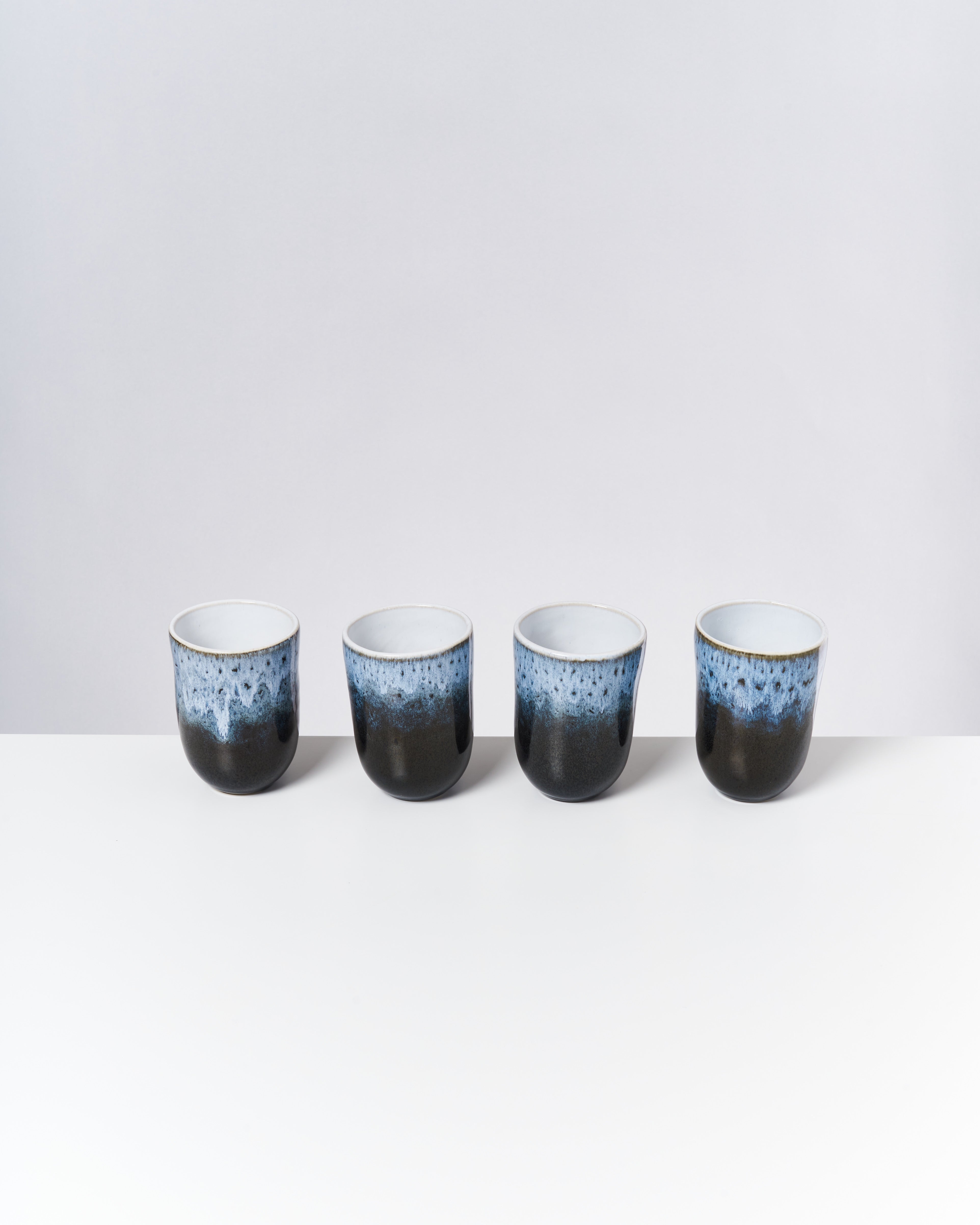 Vier schwarz-blau glasierte Keramiktassen stehen in einer Reihe auf einem weißen Tisch vor einem grauen Hintergrund.
