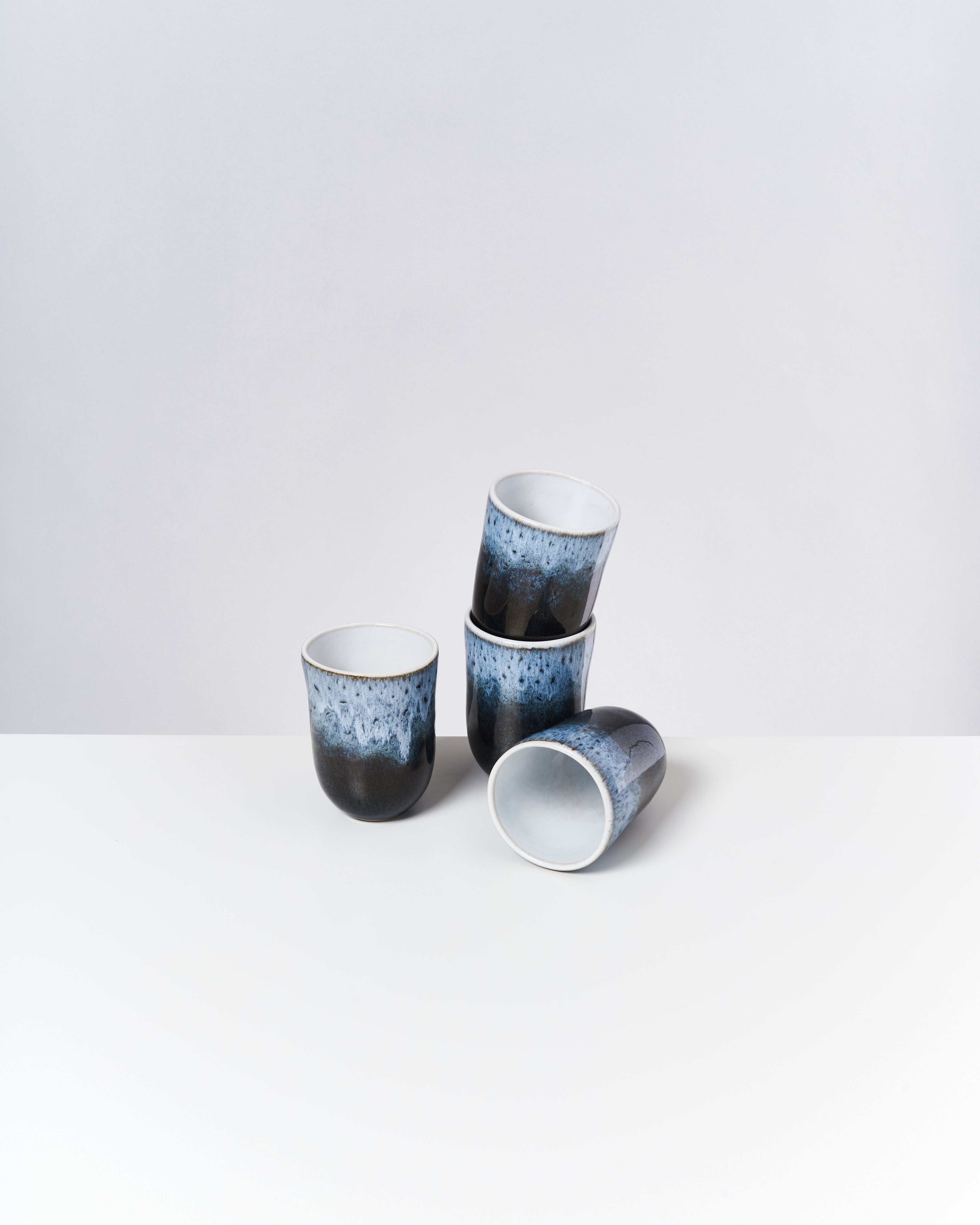 Vier blau-schwarz gemusterte Becher, von denen einer auf der Seite liegt, auf einer weißen Oberfläche.